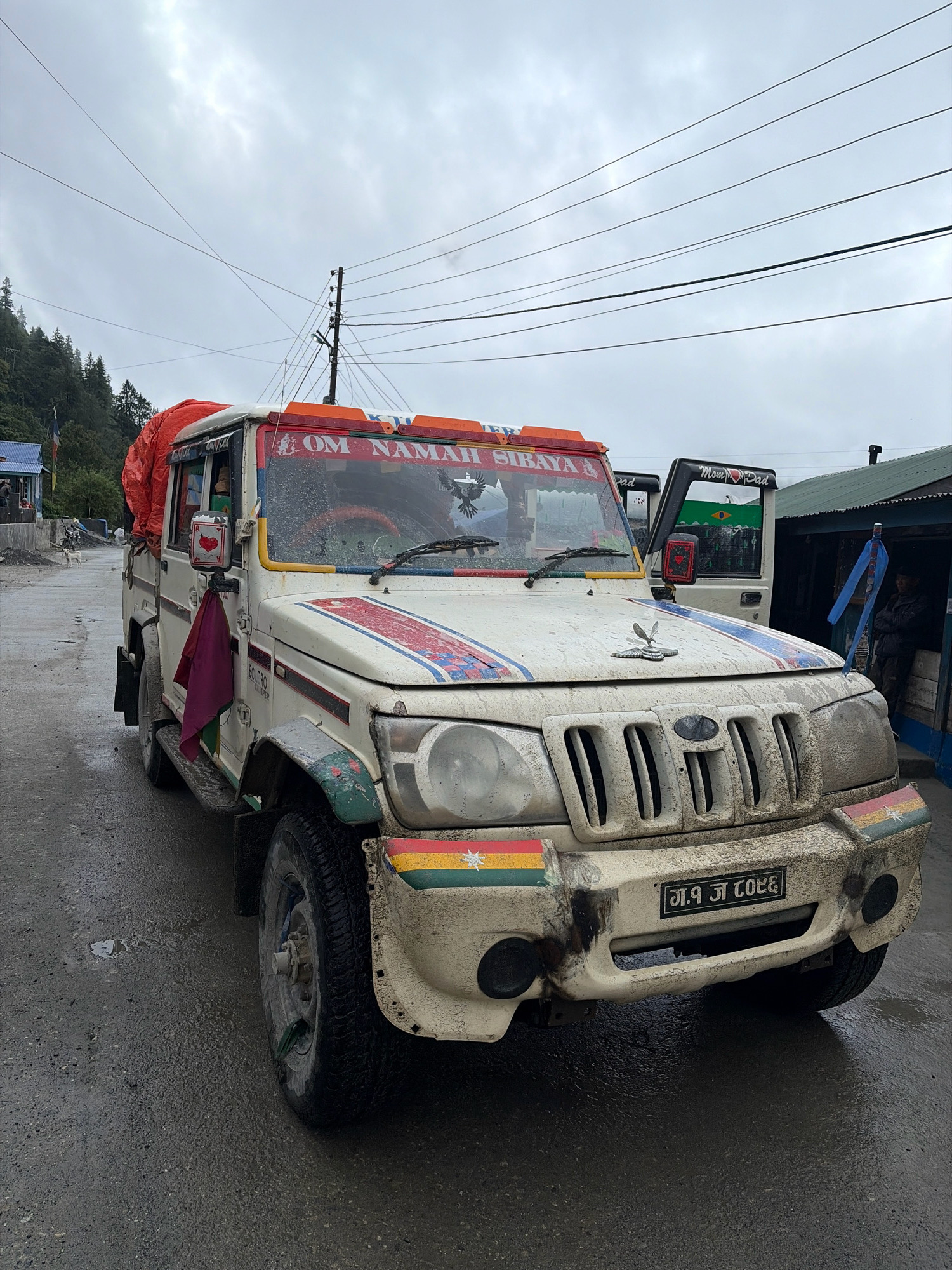 A white Mahindra Bolero, covered in mud and adorned with colorful decals, is parked on a wet, paved road in Timang, Nepal. The vehicle has a large orange tarp strapped to its roof. A red and blue stripe runs across the hood, and the front bumper features flags in red, yellow, green, and white. A license plate with Nepali script and numbers is visible on the front. A person in dark clothing stands to the right, near a building with a green roof. Another vehicle, partially visible behind the Bolero, has "Mom & Dad" written on its side. The background shows a forested hillside under a cloudy sky. A utility pole with multiple wires stands to the left of the Bolero.