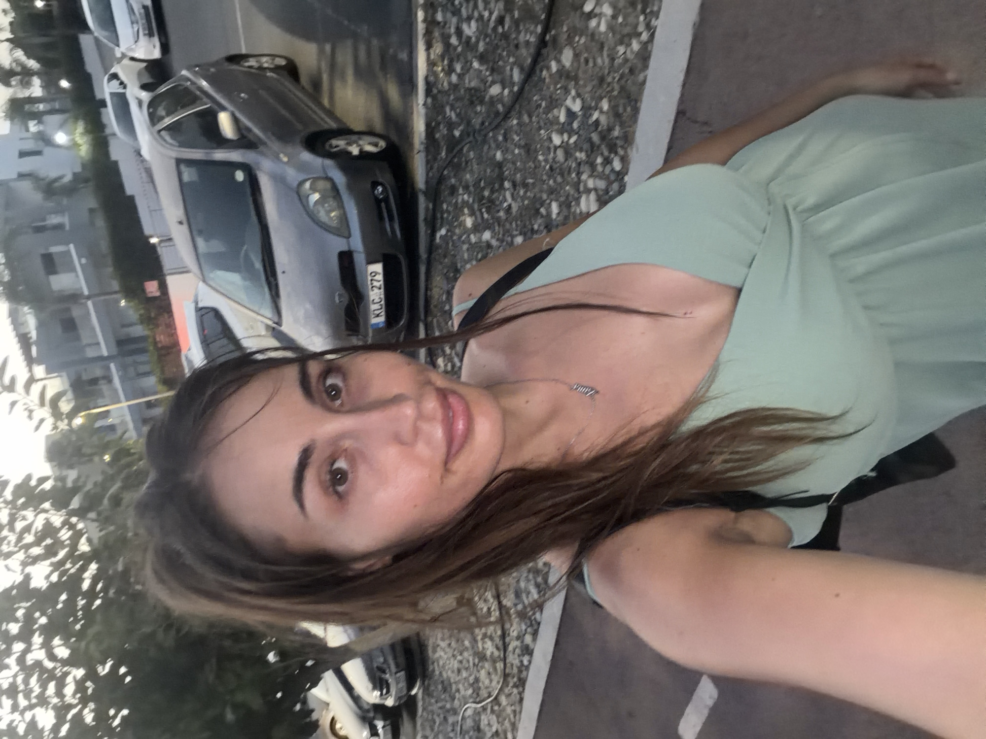 A woman with brown hair pulled into a ponytail, wearing a light green V-neck dress, is taking a selfie. She is standing outdoors in what appears to be a parking lot. Behind her, several cars are parked, including a silver Toyota Yaris with a license plate reading "KLC 279". In the background, there are buildings with balconies and some trees. The lighting suggests it is either early morning or late evening. The scene is located in Pervolia, Cyprus.