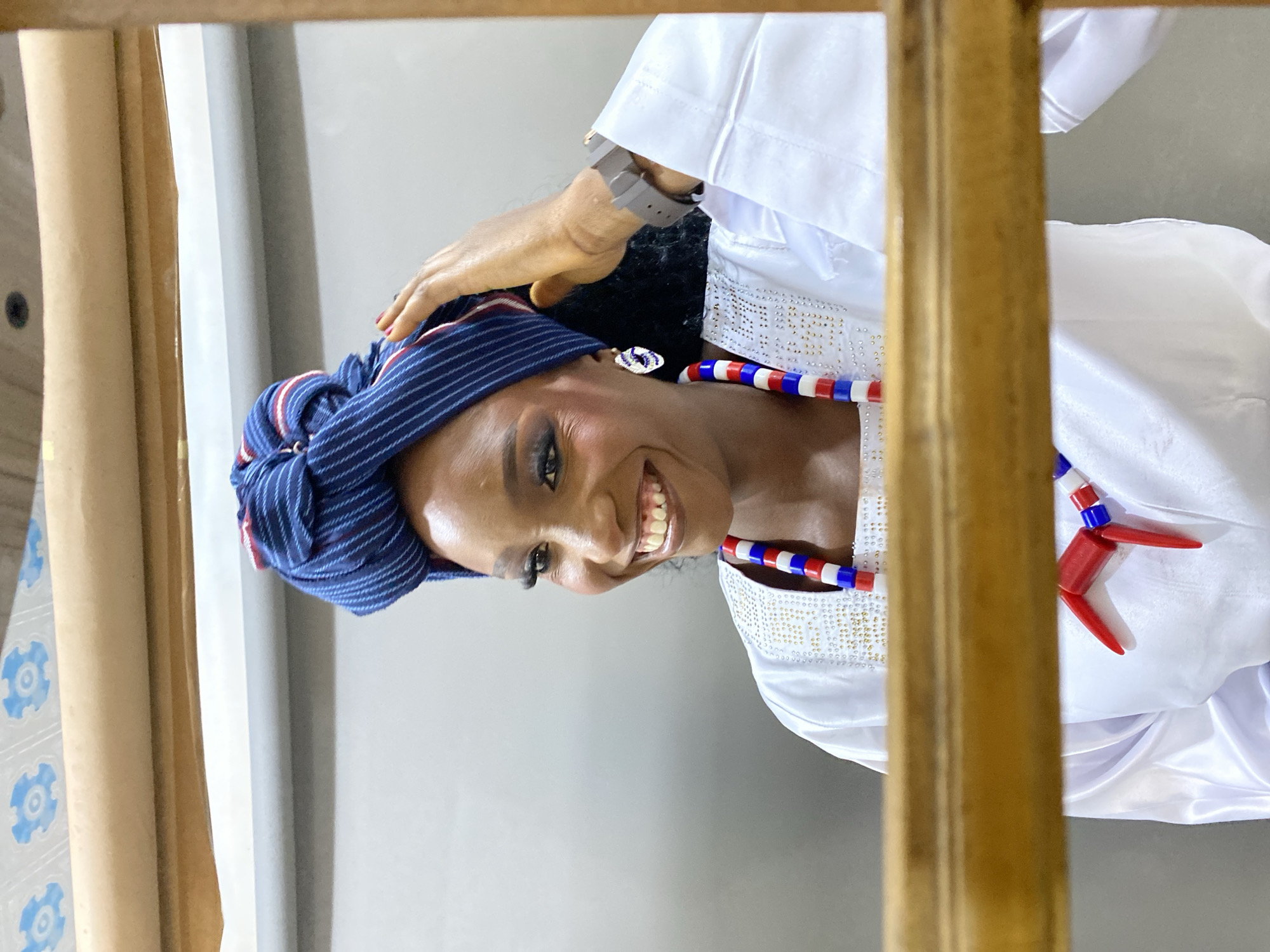 A woman, adorned in traditional white attire and a blue and white striped headwrap, smiles warmly for the camera. She wears a beaded necklace with red, white, and blue beads, and a striking red pendant in the shape of a stylized human figure. The woman's hand rests on her head, and she is wearing a grey wristwatch on her left wrist. The setting appears to be indoors, with a light-colored wall and tiled flooring visible in the background. The image is framed by a wooden element, suggesting it might be a photograph taken through a window or doorway. The woman's makeup is prominent, with defined eyes and lips, and her teeth are visible in her open-mouthed smile, conveying a sense of joy and confidence. The overall impression is of a celebratory or ceremonial occasion, possibly in Gura Top, Nigeria, given the contextual information.