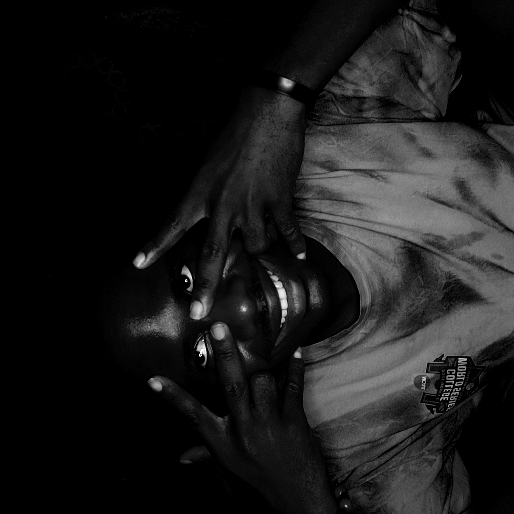 A high-contrast, black and white portrait captures a person from Narayi, Nigeria, in an expressive and playful pose. The subject has dark skin and appears to be looking upwards at the camera, possibly while lying down, as the image is rotated. They wear a patterned or tie-dye t-shirt and a simple, dark bracelet on their left wrist.
The person's hands are a central element, framing and partially obscuring their face. One hand covers the upper part of the face, with fingers spread, revealing bright, wide-open eyes that gaze directly at the viewer. The other hand is positioned near the chin, partially covering the lower jaw but clearly exposing a broad, radiant smile with prominent white teeth. The overall expression conveys joy, warmth, and a sense of playfulness or amusement.
The background is uniformly dark, creating a strong visual emphasis on the subject and enhancing the dramatic lighting. This lighting highlights the contours of the face, hands, and the brightness of the eyes and teeth. The t-shirt features a visible logo on its chest. Though somewhat obscured and rotated, discernible text includes "MIDNITE LEAGUE" and "COTTERIE," along with the letters "VCU." The scene provides no specific time of day or weather cues, given the dark, likely indoor or controlled lighting environment.