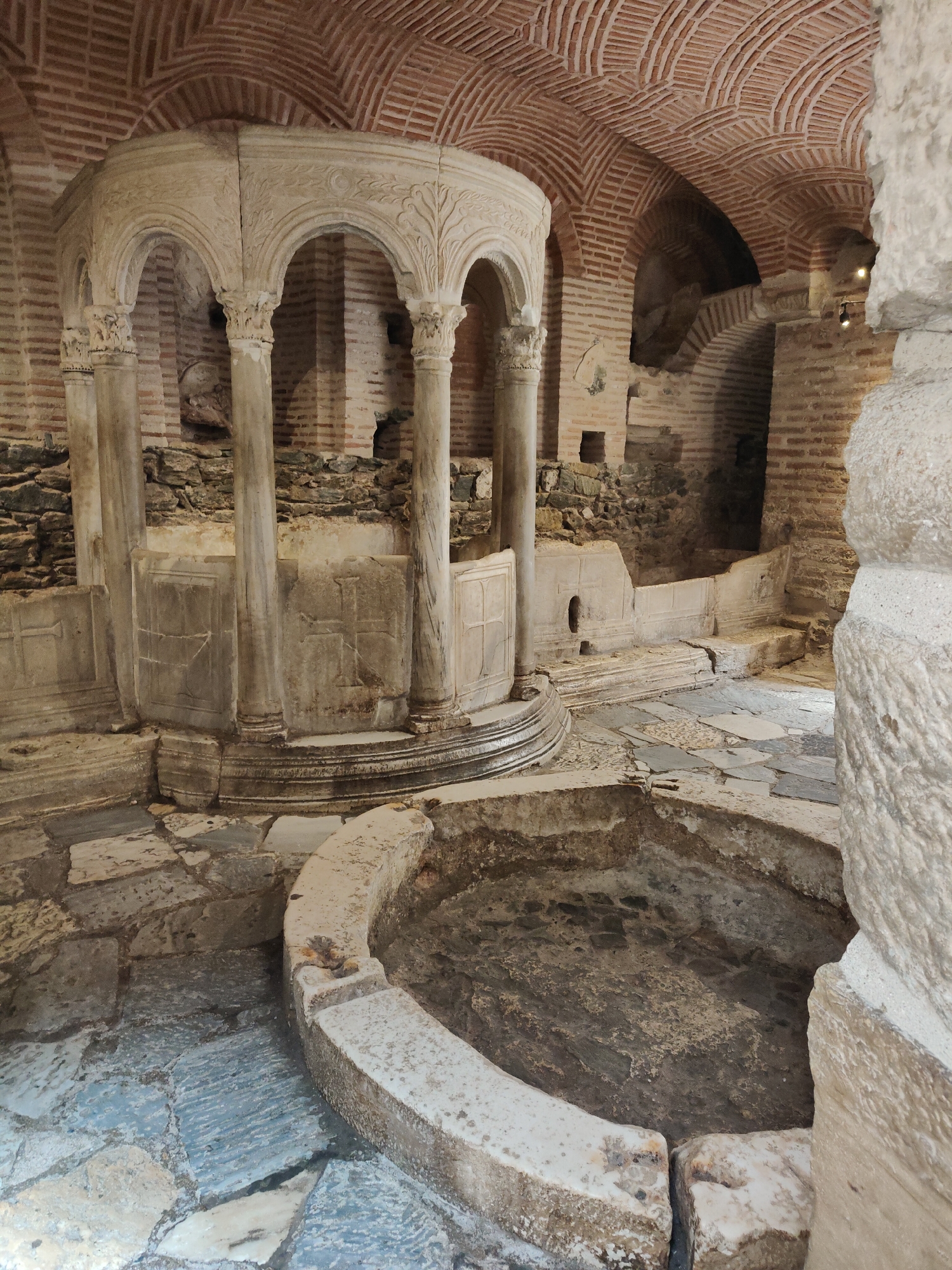 The image depicts the interior of the Rotunda of Galerius in Thessaloniki, Greece, a historic monument with a circular structure and domed ceiling constructed from brick. In the foreground, a stone basin with rough, textured walls is partially visible, its interior filled with dark, possibly water-damaged material. The floor is paved with irregular flagstones of varying colors, including grey and blue hues.
A prominent architectural element in the mid-ground is a semi-circular structure supported by ornate stone columns. These columns feature carved capitals and are topped with arches adorned with intricate floral reliefs. Behind this, the curved walls of the Rotunda are visible, built with alternating bands of brick and stone, creating a distinctive pattern. Several arched alcoves are present in the wall, some appearing dark and recessed. Scattered debris, including stones and what appears to be rubble, is present in various areas, suggesting an archaeological context. The lighting is dim, with some illumination highlighting the stone basin and parts of the floor, creating shadows that emphasize the texture and depth of the scene. No people are visible in the image.