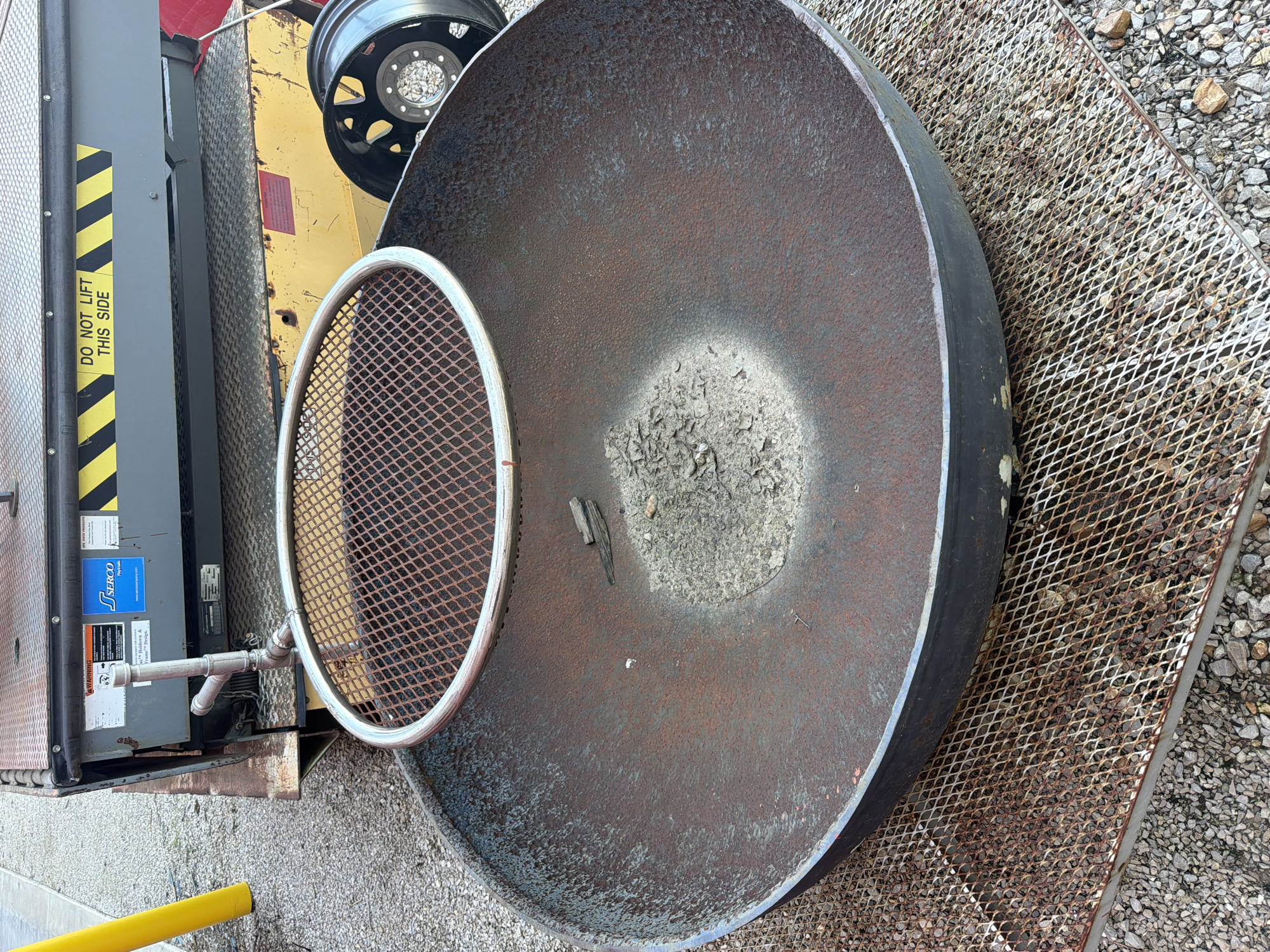 This is an outdoor, eye-level, medium shot photograph of a large, rusted metal fire pit. The fire pit is round and sits on a metal grate. In the background, there is a yellow lift gate with a "DO NOT LIFT THIS SIDE" sticker. A portion of a black tire rim is visible to the right of the fire pit. The ground surrounding the objects is covered in gravel. The scene appears to be in Hornersville, United States, though no specific landmarks are visible.
