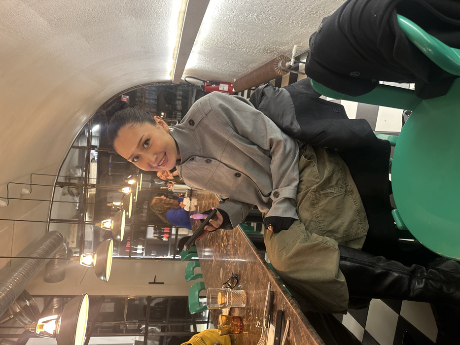 A woman with hair in a bun sits at a bar with a brown marble countertop. She wears a gray coat, green cargo pants, and black boots. She holds a cell phone in her hand and is smiling. Glasses and cutlery sit on the counter. Behind her, the bar is lit by hanging lamps, and there are other people seated. Two coats are draped over a green chair. A black and white checkered floor is visible.