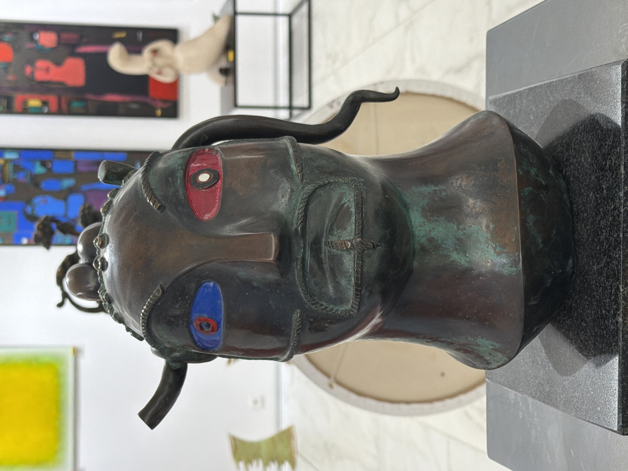 A bronze bust of a fantastical figure with a stylized face and textured skin is displayed on a dark granite pedestal in an art gallery. The figure has wide, almond-shaped eyes with striking blue and red irises. Its head is adorned with bulbous protrusions and two horns, one on each side, with a thin, coiled appendage extending from the right side of its head. The facial features are rendered with bold lines, including a prominent nose, a mustache, and a textured mouth that suggests a grimace or an open mouth with teeth. The bronze exhibits a patina, with areas of green oxidation.
In the background, blurred abstract paintings are visible on white walls. To the left, a large, light-colored circular object, possibly a canvas or drum, is partially seen. To the right, a small, light-colored sculpture of a stylized animal or figure stands on a dark metal table. The floor appears to be tiled in a light color. The lighting in the gallery is even, highlighting the sculpture and its details. The overall impression is one of a contemporary art exhibition. The information about the location in Minsk, Belarus, is not visually evident from the image itself.