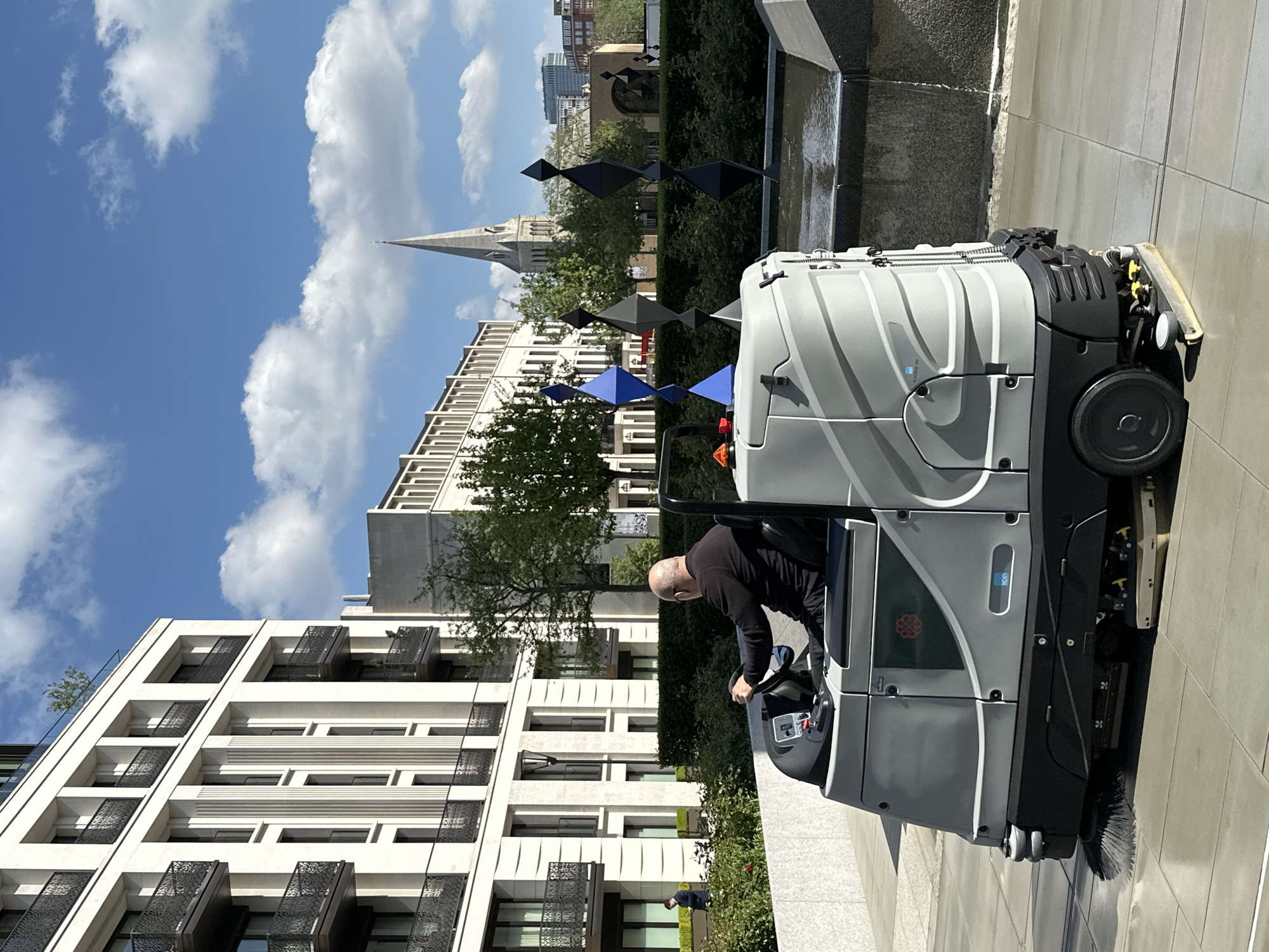 A man operates a floor cleaning machine on a paved surface in London, United Kingdom. The machine, predominantly gray with black accents and a gray-and-white striped pattern, is equipped with brushes and cleaning components. The operator, a bald man wearing a black shirt, is seated in the machine. Behind the man and the cleaning machine is a contemporary building with white facades, dark metal balconies, and evenly spaced rectangular windows. A church steeple rises in the background amidst green trees and a bright blue sky with scattered white clouds. An artwork of multiple geometric shapes is between the building and the church. Water flows from a modern stone fountain to the right.