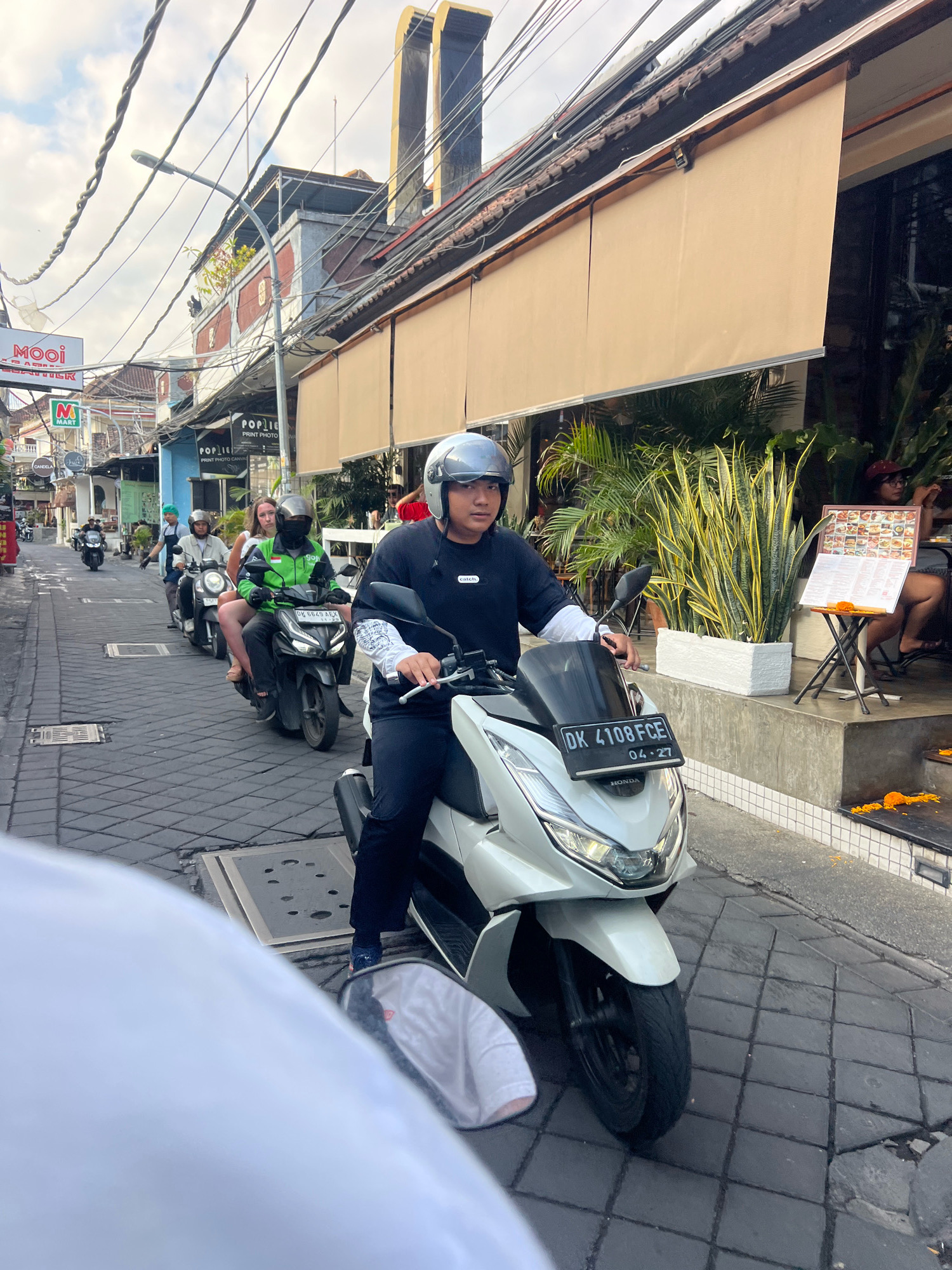 A street scene in Kuta, Indonesia, depicts a line of motorbikes on a paved street. In the foreground, a person wearing a black shirt and a silver helmet rides a white motorbike with the license plate "DK 4108 FCE." Behind them, a motorbike is ridden by a person in a green jacket, with a passenger wearing a white shirt and a black mask. Other motorbikes and riders are visible further down the street. Buildings with awnings line the right side of the street, with potted plants and outdoor seating visible. Overhead, electrical wires crisscross the sky. The overall impression is of a busy urban street scene.
