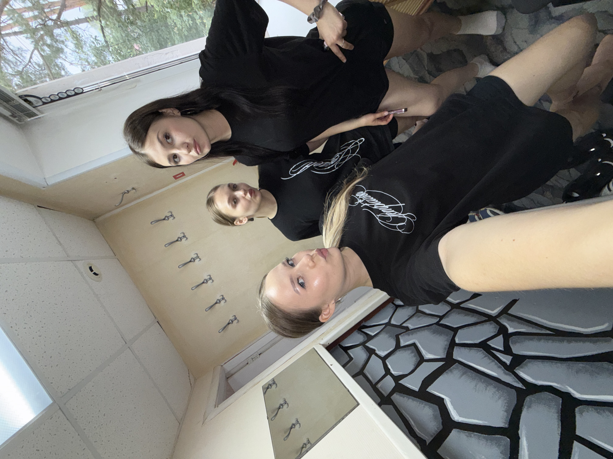 Three young women are posing for a selfie in what appears to be a room with light-colored walls and a drop ceiling. The room is decorated with what looks like a mural of large grey stones on the lower portion of one wall. Several hooks are mounted on the wall at various heights. One woman in the foreground is wearing a black t-shirt with white lettering and dark shorts. She has blonde hair pulled back and is making a kissing face towards the camera. To her right and slightly behind her is another woman with blonde hair, also wearing a black t-shirt and dark shorts, looking towards the camera with her mouth slightly open. The third woman, on the far right, has long dark hair, is wearing a black t-shirt and dark shorts, and is looking at the camera. She is holding a pink object in her right hand. A window with trees visible outside is on the right side of the image. The overall lighting is bright, suggesting daytime. The image was likely taken in the city of Chaikovsky, Russia, given the context.