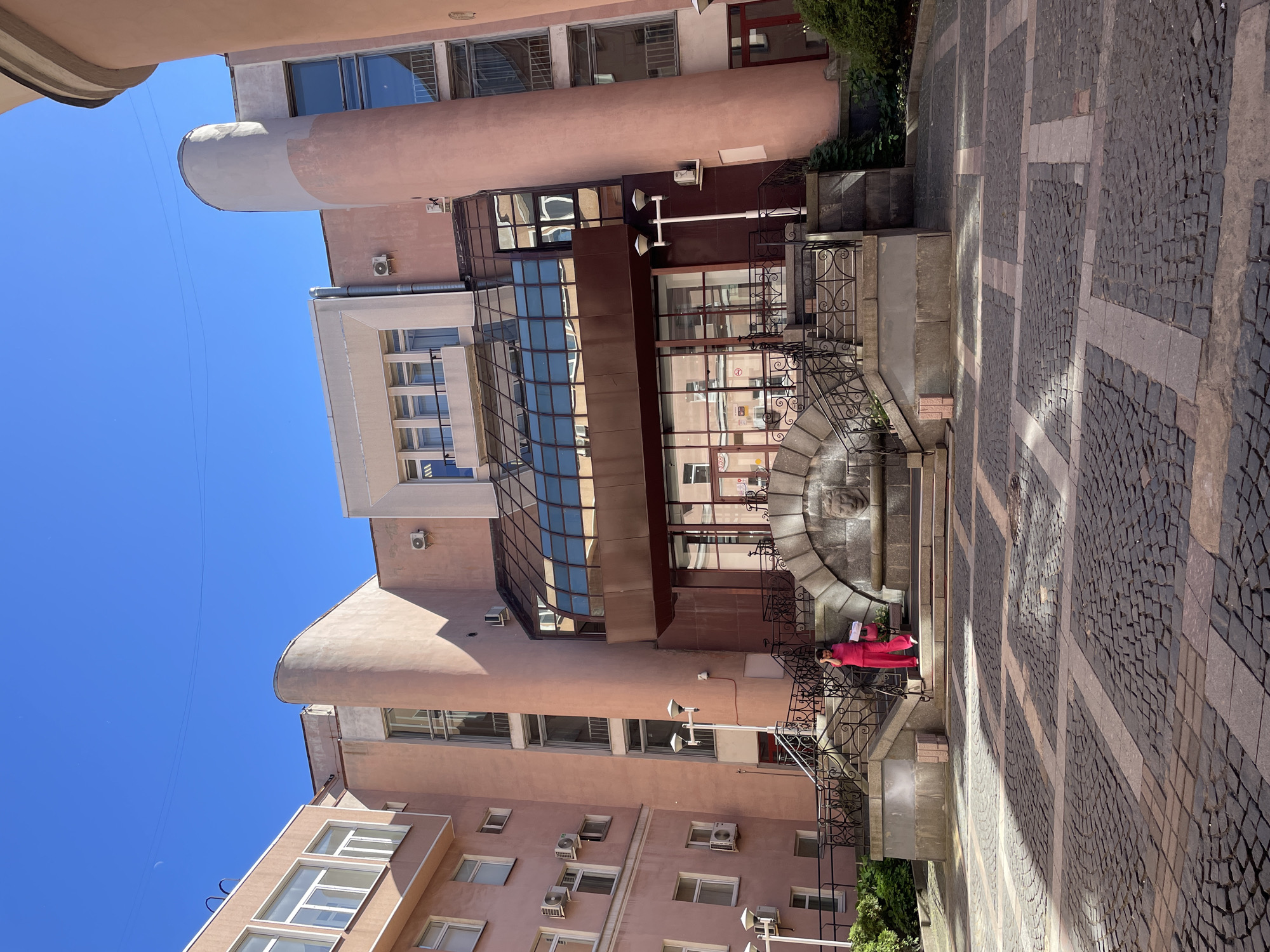 The image shows the facade of a modern, multi-story building with a pinkish-beige exterior. The building features a large, arched entrance area with a glass-enclosed canopy. A set of stairs with ornate metal railings leads up to this entrance. In front of the entrance, there is a stone fountain or decorative element with a carved lion's head.
A woman in bright pink clothing is standing on the stairs, holding what appears to be a bag or a parcel. She is facing the building and seems to be in the process of ascending or descending the stairs. The area in front of the building is paved with cobblestones in a patterned layout. The sky is clear and blue. The architectural style suggests a building located in a city, possibly an office building or a public institution. The style of the building and the paving stones are consistent with some areas in Saint Petersburg, Russia.