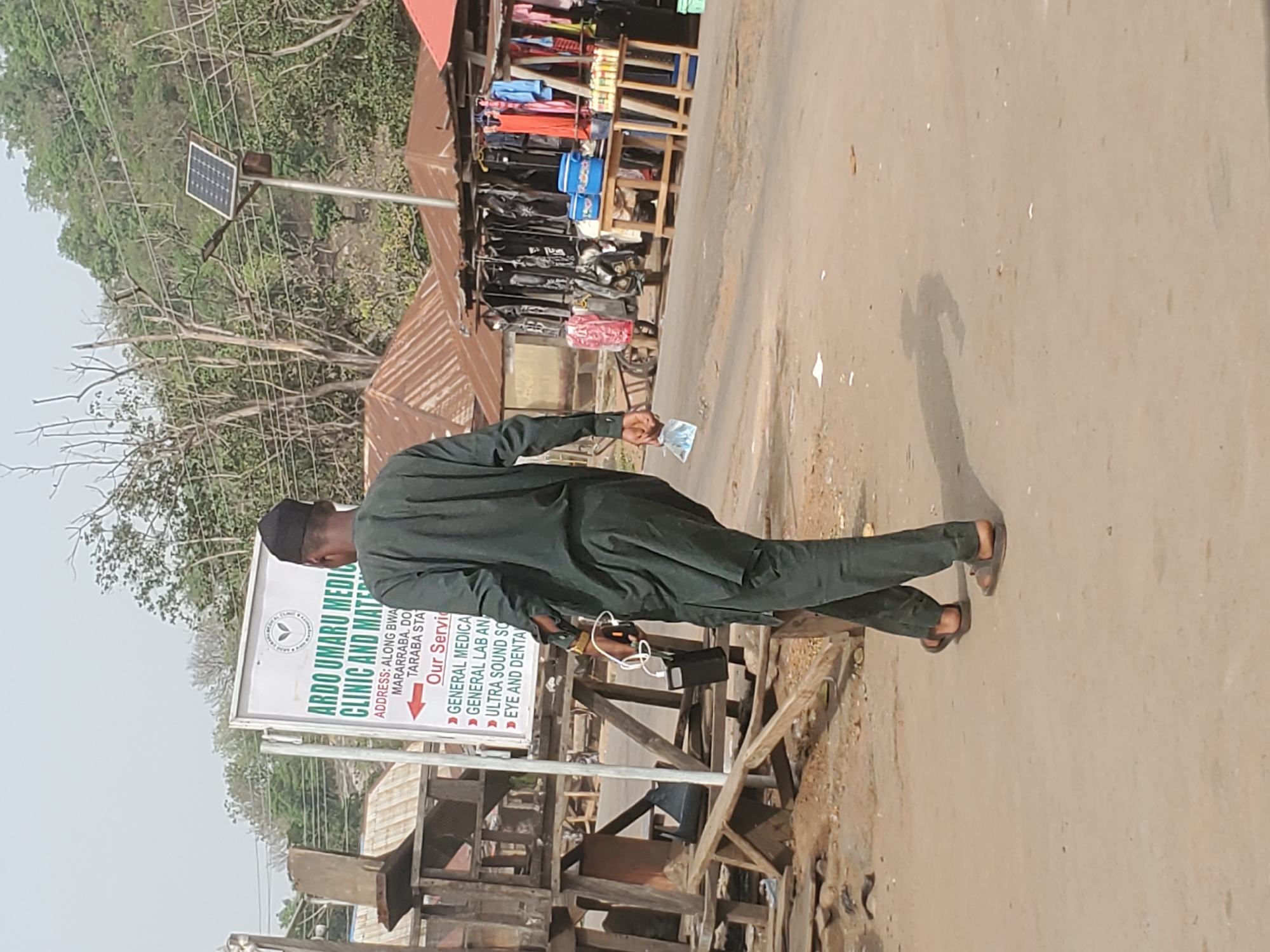 An outdoor scene captures a bright, sunny day on a street in Mararaba, Taraba State, Nigeria. A man, viewed from behind, walks along a dirt and unpaved road. He wears a dark green, possibly traditional, outfit and sandals, holding a white face mask in his right hand. His clear shadow is cast on the ground, indicating direct sunlight.
To the left of the man, a prominent white signboard advertises "ARDO UMARU MEDICAL CLINIC AND MATERNITY". The address reads "ALONG BWA MARARRABA, DO TARABA STA". The clinic's services are listed as "GENERAL MEDICAL", "GENERAL LAB AND", "ULTRA SOUND SCAN", and "EYE AND DENTAL".
Behind the signboard and further down the road, wooden structures and green vegetation are visible. On the opposite side of the road, several open-air market stalls display various goods, including clothing and bags, suggesting a bustling commercial area. A solar panel is mounted on a pole, providing power, with trees forming the background under a light sky. The overall setting appears to be a semi-urban or rural roadside with local businesses.