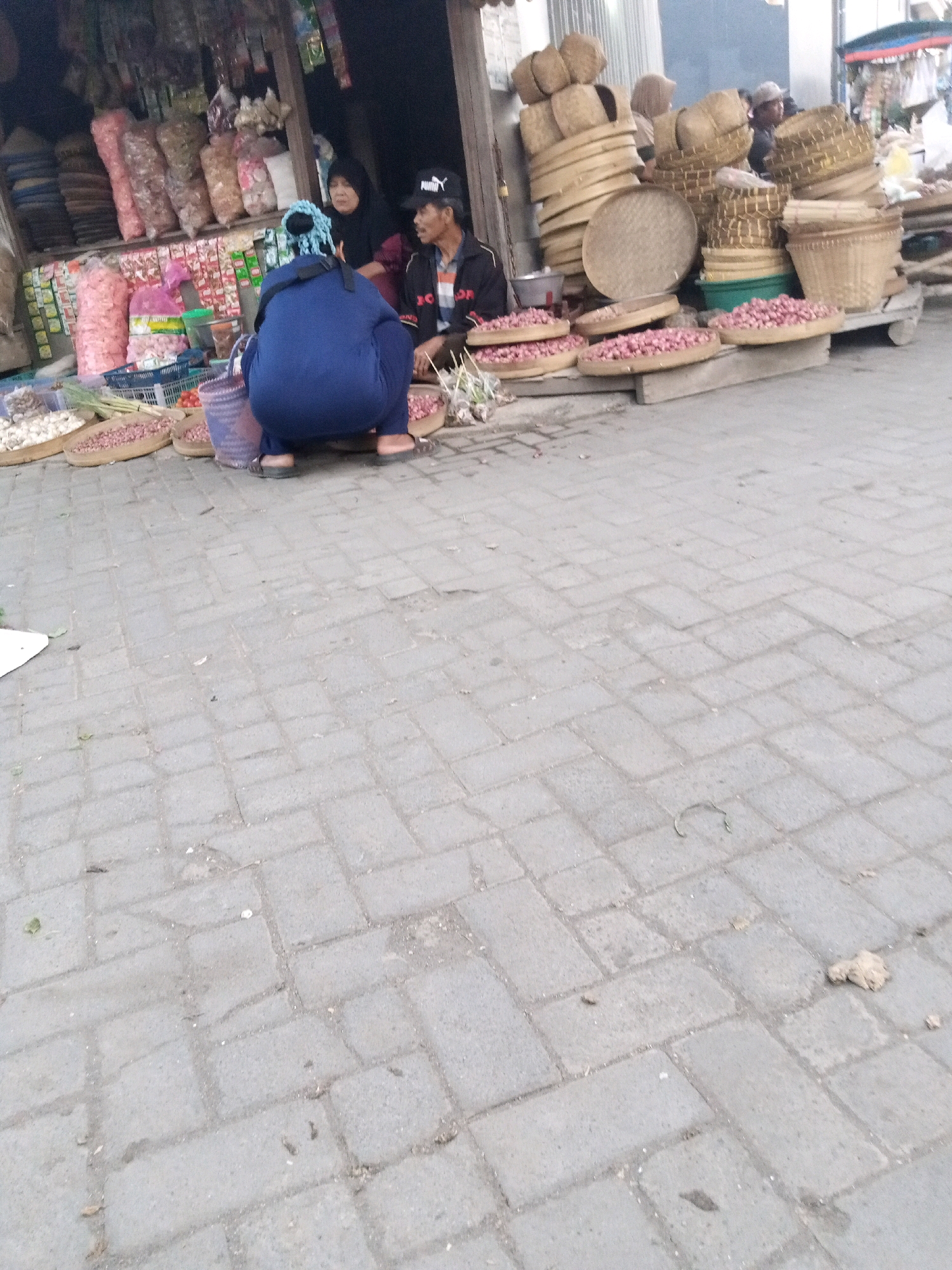 A street scene in Sono Ageng, Indonesia, shows a stall selling goods. In the foreground, a person wearing a blue outfit and headscarf squats on the ground. To their right, another person wearing a black jacket and hat is seated, facing away from the camera. Behind them, a person in a dark headscarf is also seated. The stall displays various items, including bags of produce, baskets, and what appear to be bulbs or small fruits arranged in circular trays. The ground is paved with grey interlocking bricks, and scattered debris is visible. The overall lighting suggests it is daytime.