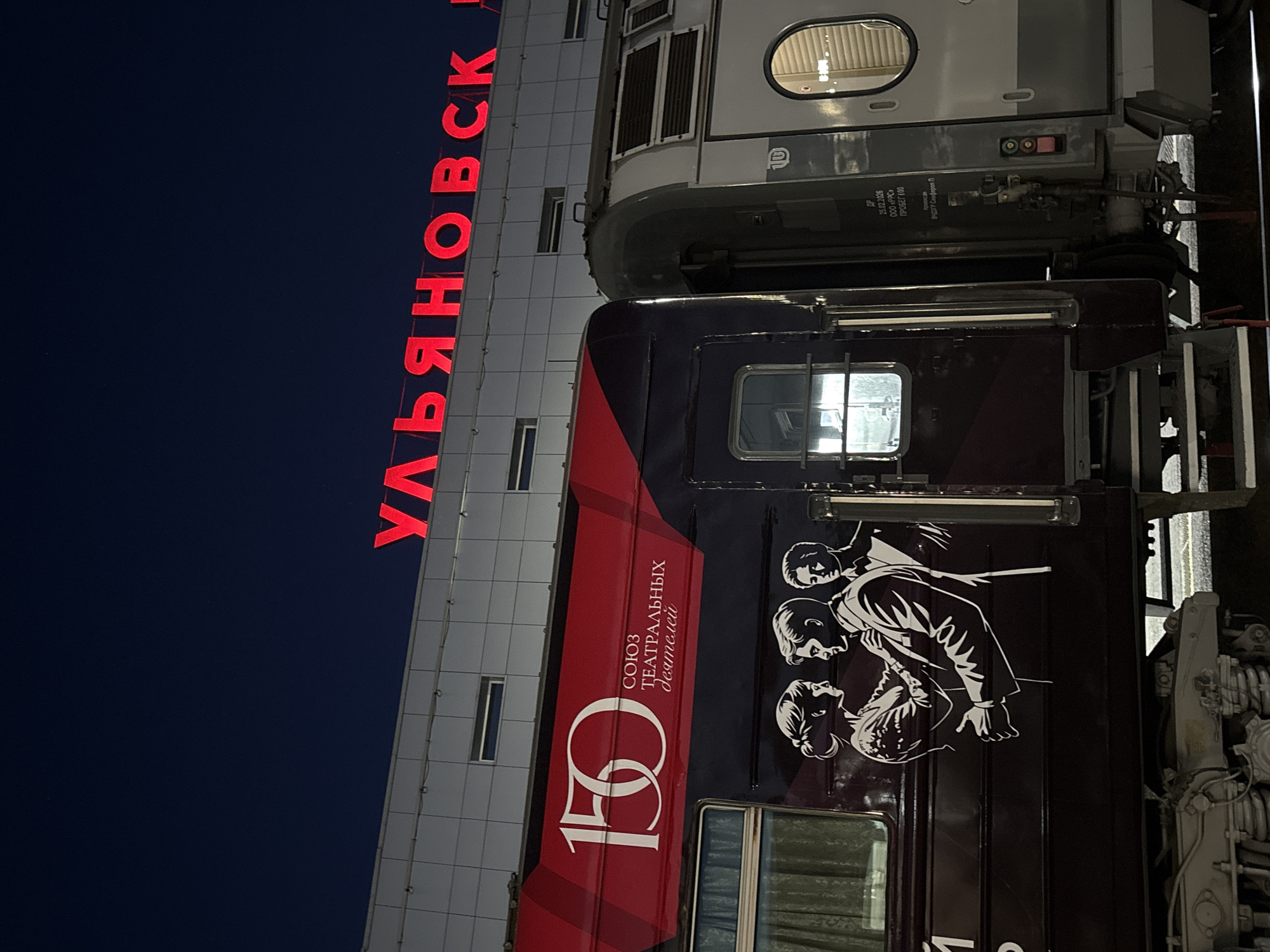 The image shows a train at a station at night, with the city's name, Ulyanovsk, displayed in large red letters on the station building. The train cars have graphic designs on them. The car in the foreground features a large "150" logo and text that reads "СОЮЗ ТЕАТРАЛЬНЫХ деятелей" which translates to "Union of Theatre Workers." Below this text is a black and white illustration of three people, two in the foreground embracing and one in the background. The other train car visible is a passenger car, gray and white, with windows. The sky is dark, indicating it is nighttime. The station building is modern with horizontal siding and rectangular windows.