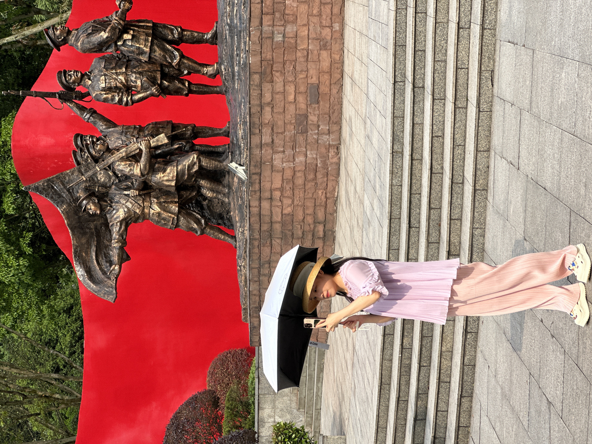 A woman with long black hair and wearing a straw hat, a light purple dress, and light pink pleated pants is standing on a stone-tiled area next to a set of concrete stairs and using her smartphone under a black and white umbrella. Behind her is a large bronze monument depicting soldiers with weapons and a flag on a red background. The monument is attached to a brick-like wall. Shrubs are visible at the base of the monument. This scene is in Liangjiang, China.