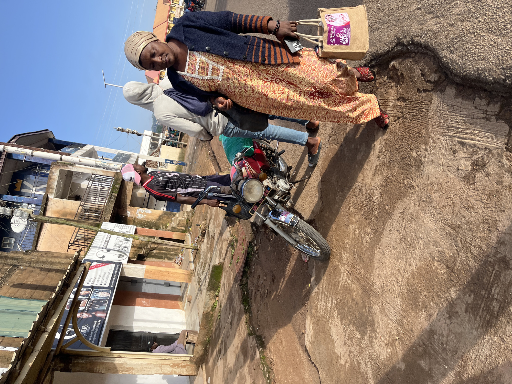 In Gura Top, Nigeria, a street scene unfolds under a clear blue sky. The foreground is dominated by a rough, reddish-brown concrete surface, marked by cracks and patches of dirt. A motorcycle, primarily red with chrome accents, stands angled on the right side of the image, its front wheel visible. To the right of the motorcycle, a woman walks towards the viewer. She wears a light-colored headwrap, a dark blue cardigan with striped sleeves, and a vibrant orange and yellow patterned dress. She carries a beige tote bag in her right hand, which features a pink and purple graphic with text that reads "FOREVER entwined ALEX & ROTMWA 5TH NOV. 2025". Her left hand holds a dark-colored smartphone. Her feet are adorned with red sandals.
Behind the woman and to her left, a man stands facing left, wearing a black and white striped t-shirt with red accents and a pink baseball cap. He appears to be looking at something off-frame. Further to the left, a person in a light grey hooded jacket and blue jeans stands with their back to the camera, a dark blue bag slung over their right shoulder. In the background, a building with multiple stories features a mix of weathered brown and blue facades, balconies with metal railings, and what appears to be a satellite dish. A sign with images of faces and text is visible on the lower part of the building. A man in a grey shirt and dark trousers is seated on a low wooden bench outside this building on the far left. The street is lined with buildings and shows signs of everyday life, with some debris visible on the ground.
