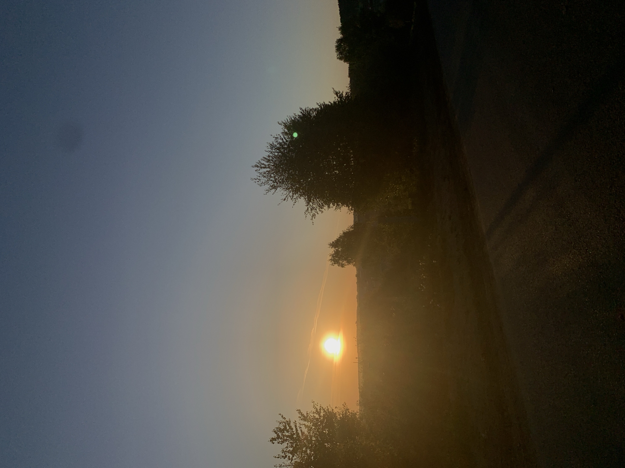 The image depicts a rural scene at sunset. The sun is low on the horizon, casting a warm orange glow. Silhouetted trees line the right side of the frame, and what appears to be a paved surface runs along the edge. The sky transitions from dark blue at the top to a lighter orange near the horizon. The image suggests a location near Фоки, Russia, characterized by a landscape where nature and infrastructure intersect. There are no people visible in the image.