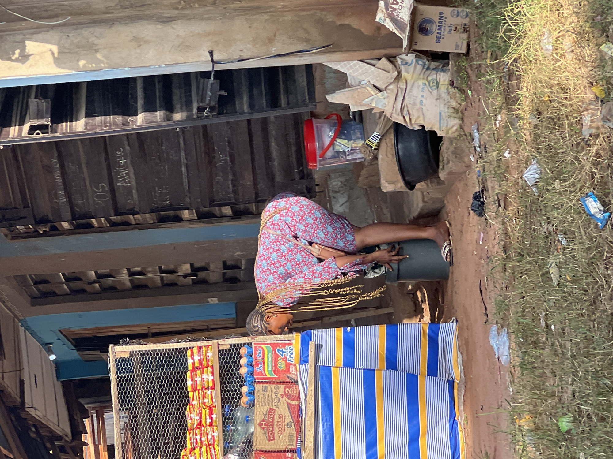 In Nkpor, Nigeria, a woman with braided hair, wearing a floral-print dress, is bent over a large grey bucket in front of a market stall. The stall displays various packaged goods, including noodles and eggs. The ground is dirt and covered with dry grass and scattered litter. To the right of the woman, there are several sacks, a black basin, and a box labeled "SEAMANS HAIR GRO." In the background, a wooden structure with a blue painted section and dark wooden shutters is visible. The overall scene depicts a typical informal market setting.