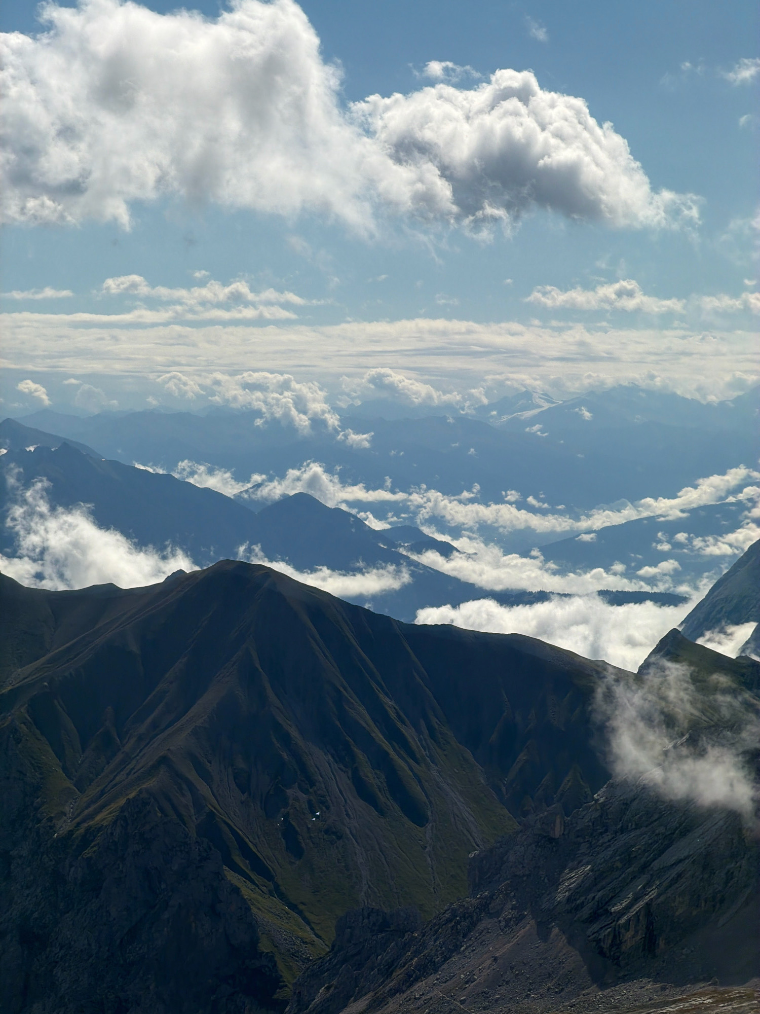 This is a high-angle, vertical, medium shot of a mountain range with clouds in the sky. The foreground features dark, rugged mountain slopes covered in patches of green vegetation. Further in the distance, successive layers of mountains recede into a hazy blue atmosphere, interspersed with pockets of white clouds. The sky above is a light blue, dominated by large, fluffy white cumulus clouds, with a few thinner clouds stretching towards the horizon. There are no people or discernible man-made objects in the image. The scene depicts a natural mountainous environment.