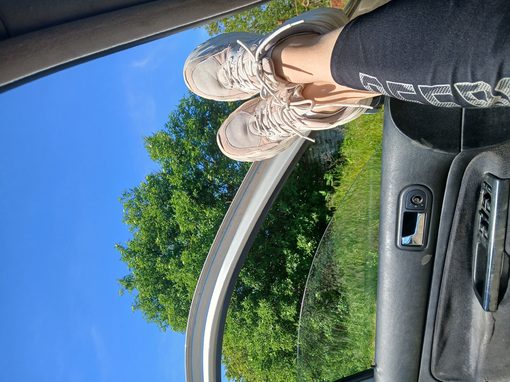 The photograph depicts a person's legs resting on the open door of a car, presumably while parked in a natural setting. The person is wearing black leggings with white text on the lower leg and light-colored sneakers. The legs extend from inside the car, resting on the upper door frame. The car's interior door panel is visible with a handle and window controls. Outside the car, there is a lush green tree against a clear blue sky. The location is claimed to be Sokolovo, Bulgaria. However, there are no landmarks or indicators to definitively confirm this geographic location. The composition is rotated.