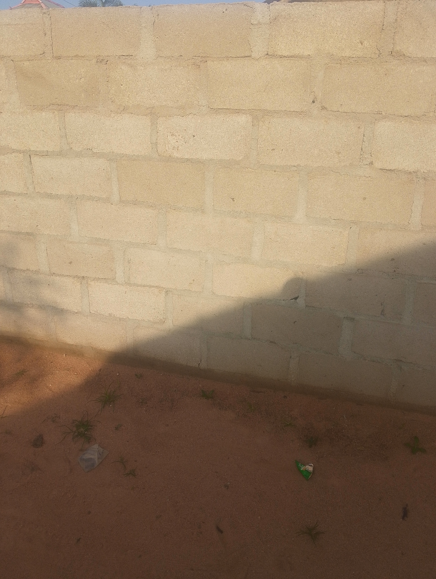 The image depicts a section of a cinder block wall, painted a light tan color, possibly in a construction or undeveloped area. The wall is constructed from rectangular blocks laid in a staggered pattern, with visible mortar lines. Sunlight casts a shadow diagonally across the lower portion of the wall. The ground in front of the wall is composed of reddish-brown sand with sparse patches of dry grass. A crumpled plastic wrapper, light-colored with some blue markings, lies on the sand to the left of the shadow. Further to the right, another small, green and white piece of litter is visible on the sand, possibly a candy wrapper or a sachet. The top edge of the image reveals a sliver of a building with a red roof and some green foliage, suggesting an outdoor setting near other structures and vegetation. The overall scene suggests an area that may be undergoing construction or is a less maintained part of a town or city. Given the location of Jalingo, Nigeria, this scene could represent a typical environment found in residential or developing areas within the city.