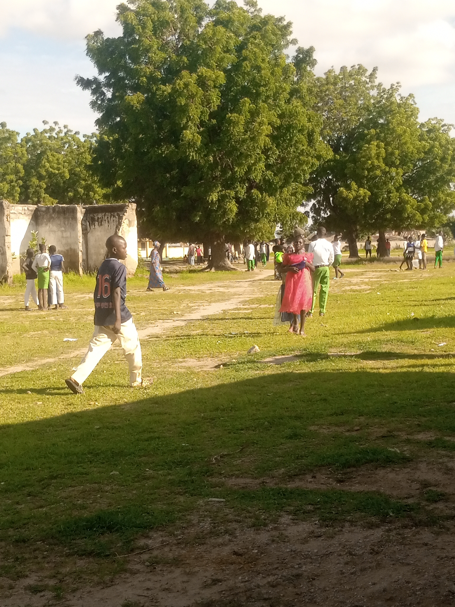 A group of people are gathered in a grassy outdoor area with some dilapidated structures in the background. A young boy in the foreground is walking towards the right of the frame, wearing a dark blue t-shirt with the number "16" on the back and light-colored pants. To his left, a few people, possibly students, are standing near a crumbling wall; they are wearing various colored clothing, including a white top and green pants. Further in the background, a woman in a blue and white patterned dress is walking. To the right, a woman in a bright pink dress is standing with a person in a white shirt and green pants behind her. Several other individuals and groups of people are scattered throughout the scene, some standing, some walking. The environment appears to be a somewhat neglected public space, with large trees providing shade and patches of dry earth mixed with grass. The sky is visible and appears partly cloudy. The overall impression is of a casual gathering or a scene in a community area. The location is identified as Langtang, Nigeria.