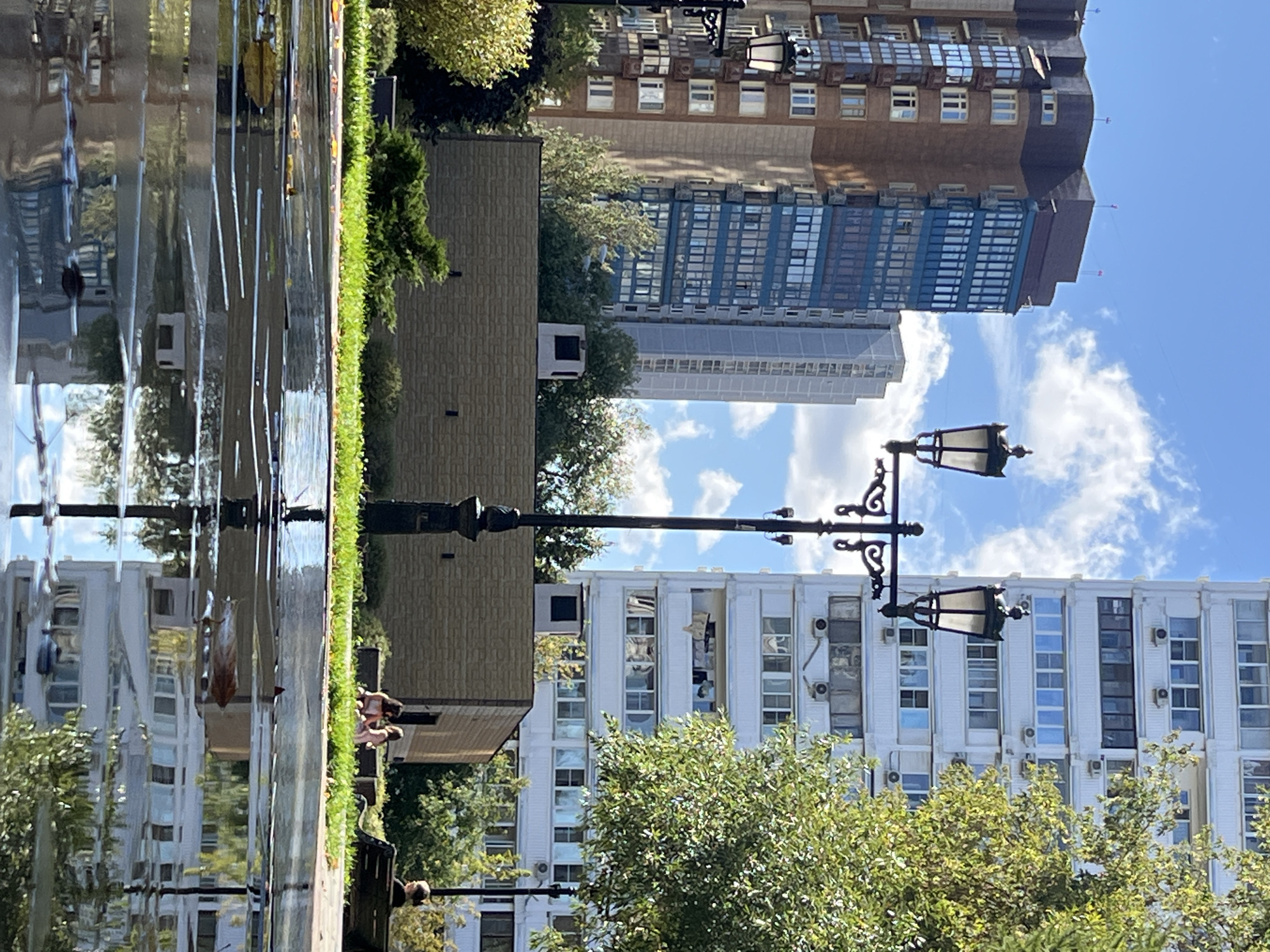 The image depicts an urban scene in Moscow, Russia, on a bright, sunny day with scattered clouds. In the foreground, a wet pavement surface reflects the surrounding environment, including the sky, trees, and buildings. Several people are visible in the mid-ground, including two individuals sitting on the grass and one person on a park bench.
The background is dominated by multi-story residential buildings. To the left, a taller, modern building features a brown facade with a prominent blue glass section. To the right, a white, monolithic apartment building with numerous windows and air conditioning units is visible. A lamppost with multiple lights stands prominently in the middle of the frame. Lush green trees and manicured grass areas contribute to the park-like setting. The overall atmosphere is one of a typical city day, with residents enjoying the outdoors in a well-maintained urban space.