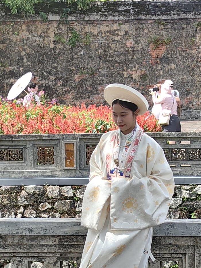 The scene is set outdoors in Phú Xuân Ward, Huế, Vietnam, likely within a historical complex such as the Imperial City. In the foreground, a young East Asian female stands centrally, facing slightly to her left. She wears a wide-brimmed, flat white traditional Vietnamese hat and an elaborate cream-colored long-sleeved robe with gold floral patterns and a complex multicolored embroidered collar. A multi-strand pearl necklace is draped around her neck. Her hands are clasped or folded at her waist, and her gaze is directed downward.
Behind her, a grey stone balustrade with ornate square cutouts extends horizontally. Beyond this balustrade, a dense bed of red flowering plants forms a vibrant mid-ground. To the left, partially obscured by these flowers, a second female in light-colored attire holds an open white parasol. To the right, a third female, dressed in a pink top, dark pants, and a white cap, is actively holding a smartphone or camera at eye level, appearing to photograph something beyond the frame. A fourth individual is partially visible behind her. The background consists of a tall, weathered brick wall, exhibiting discoloration and sparse green vegetation. Below the balustrade, a rough stone wall foundation is visible. The scene is brightly lit under direct daylight.