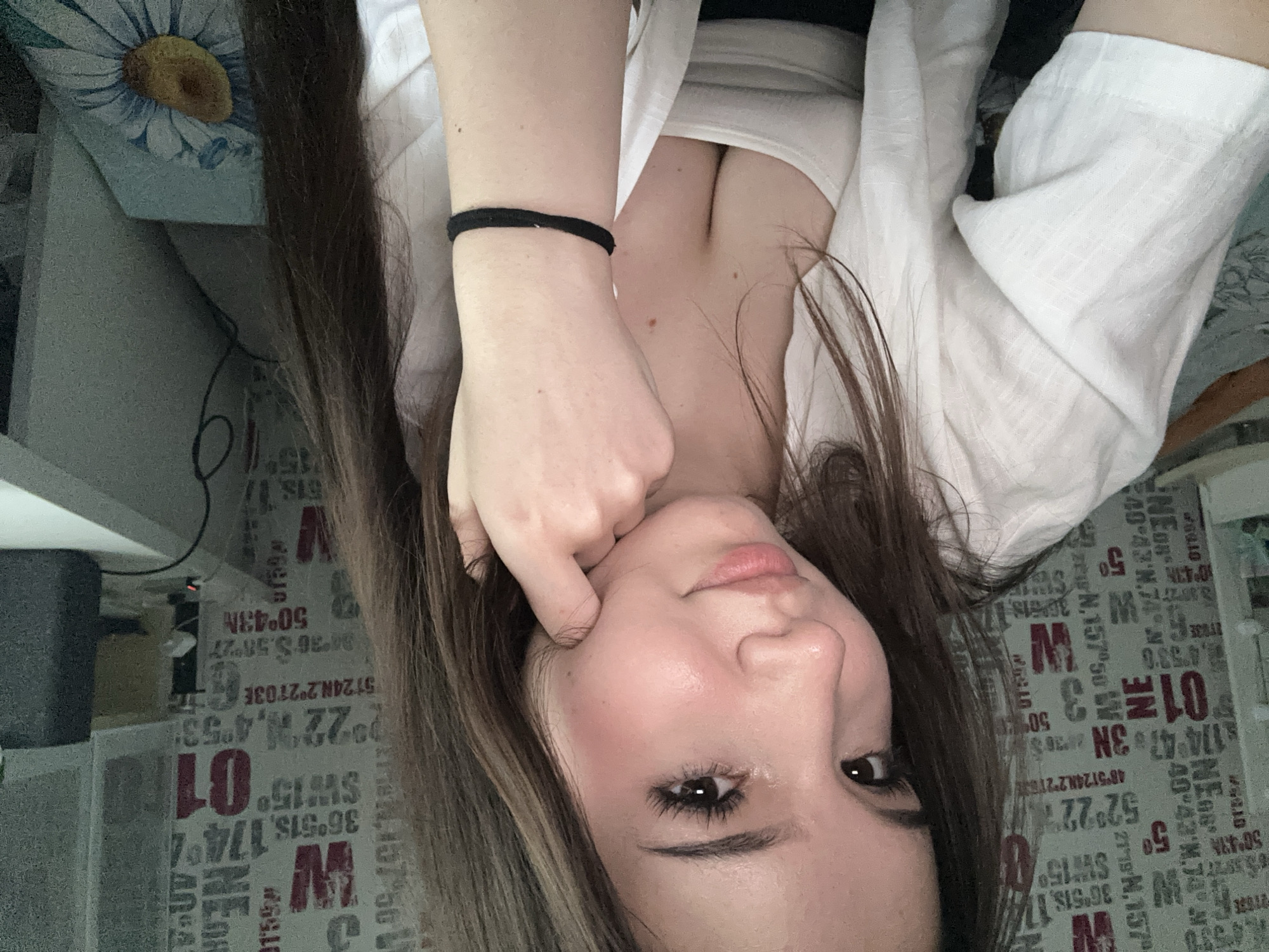 The image is a medium shot of a young woman with long, dark brown hair. She is wearing a white V-neck top underneath an open white button-down shirt. Her right hand is raised, with her fingers resting on her cheek. She is looking directly at the camera with a slight smile. The background appears to be a room with patterned wallpaper and some objects on a shelf, including what looks like a speaker. The lighting is soft, suggesting it may be indoors during the daytime or with artificial lighting. The location is not identifiable as Naberezhnye Chelny, Russia based solely on the visual information provided.