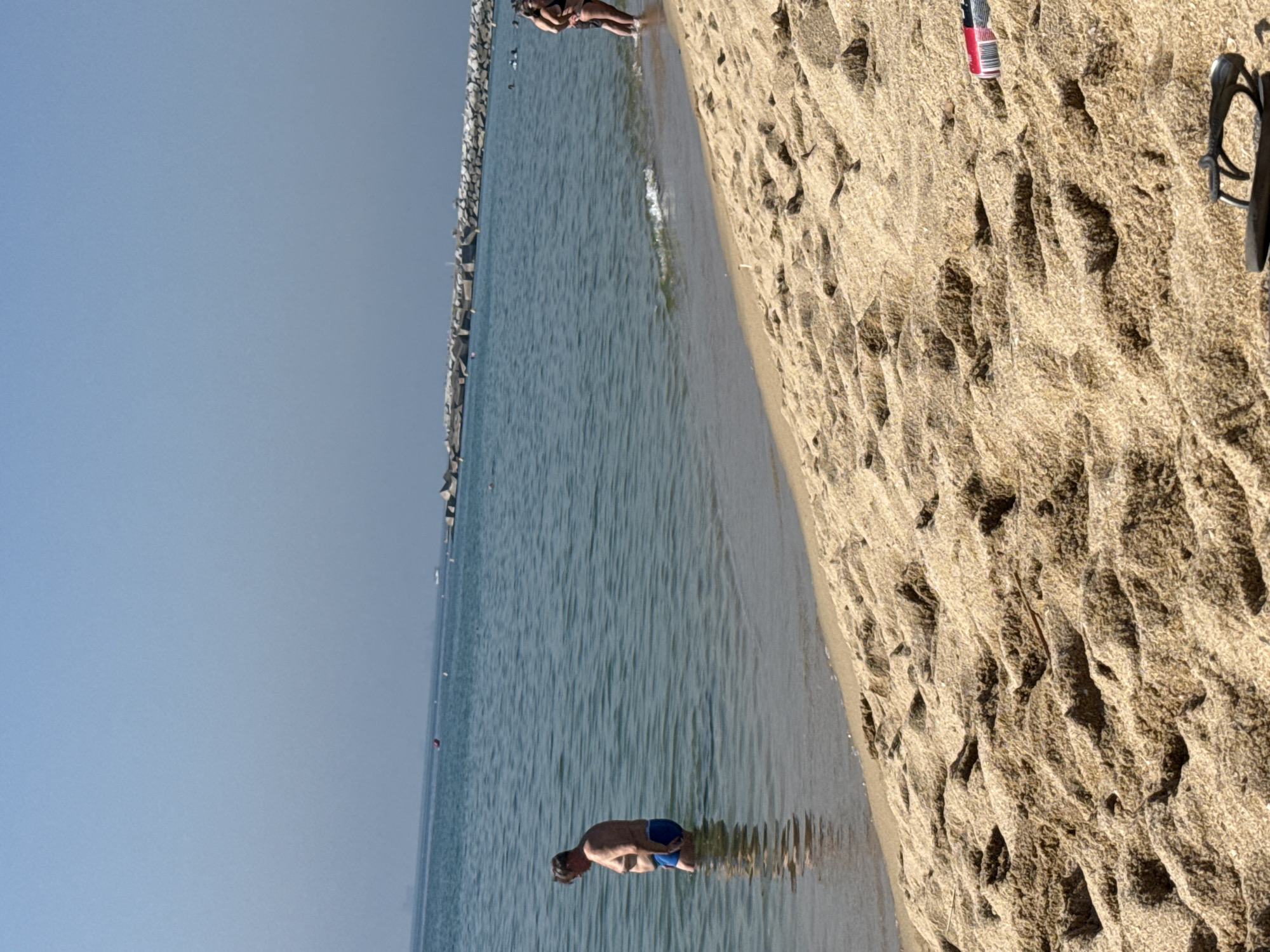 This image depicts a sunny day at a beach in Barcelona, Spain. In the foreground, the sandy beach is textured with footprints. Further back, the shallow, blue water laps at the shore. A man in blue swim trunks stands in the water, facing away from the camera. In the distance, a breakwater made of large rocks extends into the sea. A couple is visible standing at the water's edge further down the beach. The sky is clear and bright blue.