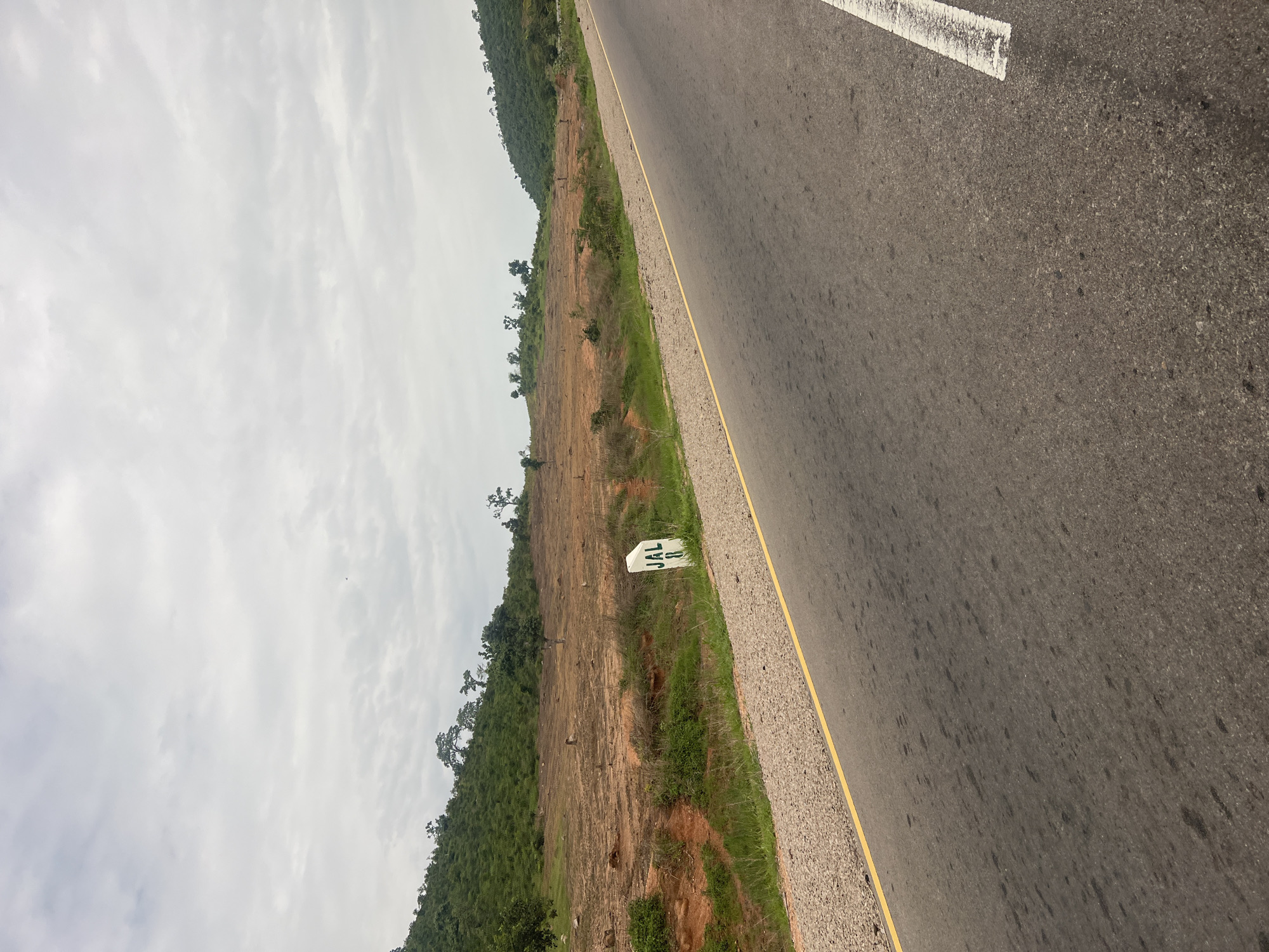 The image shows a roadside scene in Minda, Nigeria. The focus is on a highway with a gray asphalt surface and a yellow line marking the edge of the lane. A white concrete marker with green lettering, "JAL 8", is visible on the grassy verge beside the road. The land slopes upwards from the road, covered in dry earth, sparse green grass, and scattered trees. Further up the slope, the vegetation becomes denser, forming a green hillside. The sky is overcast with white and gray clouds, suggesting it is daytime. The overall impression is of a rural or semi-rural highway environment under cloudy skies. There are no people or vehicles visible in the image, and the scene appears quiet and still.