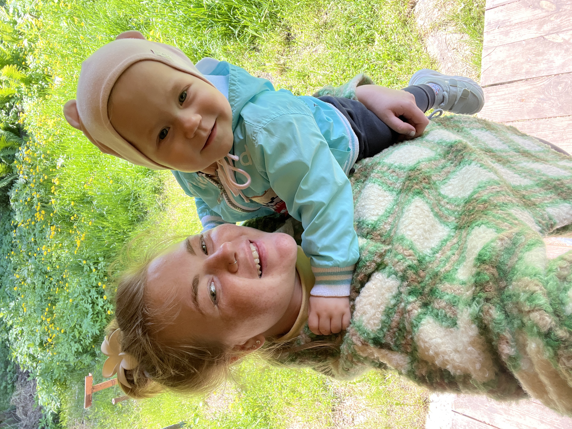 A woman and a toddler are lying on a blanket outdoors. The woman, with fair skin and blonde hair styled with a flower, is on her back. The child, wearing a pink hat and a light blue jacket, is lying on the woman's stomach. The background shows green grass and foliage, including yellow flowers. A wooden surface is visible in the top right corner. This is likely a candid, personal photograph taken in an outdoor setting in the city of Chaikovsky, Russia.
