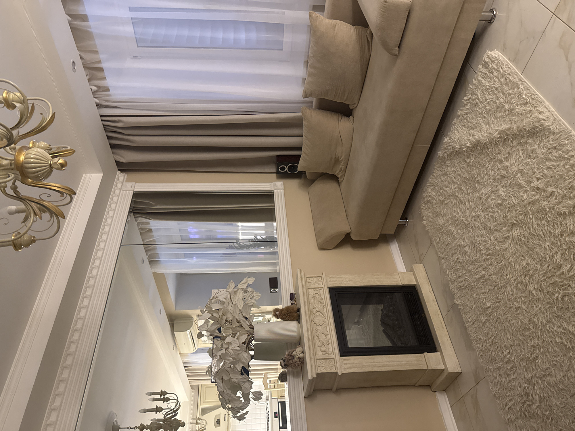This is an indoor photograph of a living room. The room has a light-colored, possibly beige or cream, sofa with two large throw pillows. In front of the sofa is a fluffy, cream-colored rug. To the left of the sofa is a decorative fireplace with a faux fireplace insert, topped with a vase filled with white artificial flowers and two small stuffed animal figurines. A large mirror spans the wall above the fireplace, reflecting part of the room and what appears to be a kitchen area in the background. The room is decorated with elegant molding along the ceiling and a chandelier hangs from the ceiling. There are beige curtains covering a window on the right side of the image, with sheer white curtains layered in front of them. The walls are painted a pale yellow. The floor is tiled with a marble-like pattern. The lighting suggests it is evening or the room is dimly lit. The image does not contain any information that would indicate the location is Moscow, Russia.