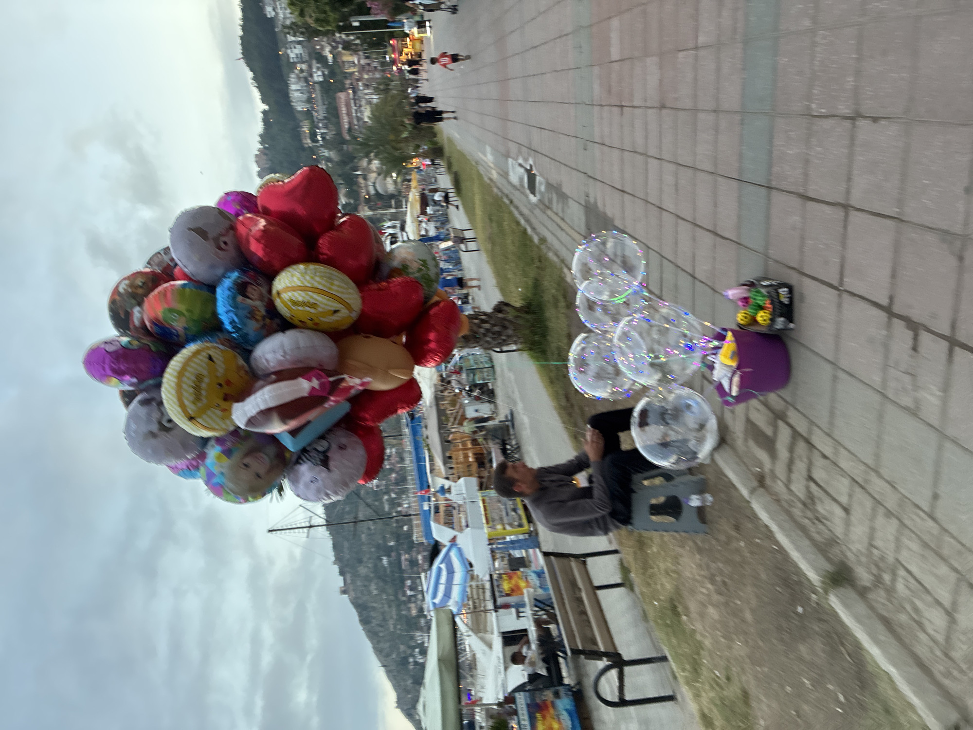 The image captures a twilight scene along a paved promenade in Alanya, Turkey. In the foreground, a person, likely a vendor, is seated on a bench, facing a display of balloons. To their left, a large cluster of colorful foil balloons floats, including red heart-shaped balloons and various circular ones. Some circular balloons feature cartoon characters or designs, such as a yellow one resembling Pikachu and another with text possibly reading "Happy Birthday." To the vendor's right, several clear, bubble-like balloons are visible, illuminated from within with colorful LED lights. Next to these clear balloons, a small black box or stand sits on a purple container.
The paved walkway extends into the background, where other people can be seen strolling. A narrow strip of grass separates the walkway from further establishments and structures. Beyond this, a town with buildings nestled on a hillside rises, and what appears to be a marina or waterfront with boats is visible. The sky is overcast, contributing to the soft, diffused light, suggesting it is either dusk or early evening, which is further implied by the glowing lights within the clear balloons. The overall atmosphere appears calm, indicative of an evening leisure activity. No explicit text is clearly legible on any signs or distant objects, apart from the possible partial text on the balloons.