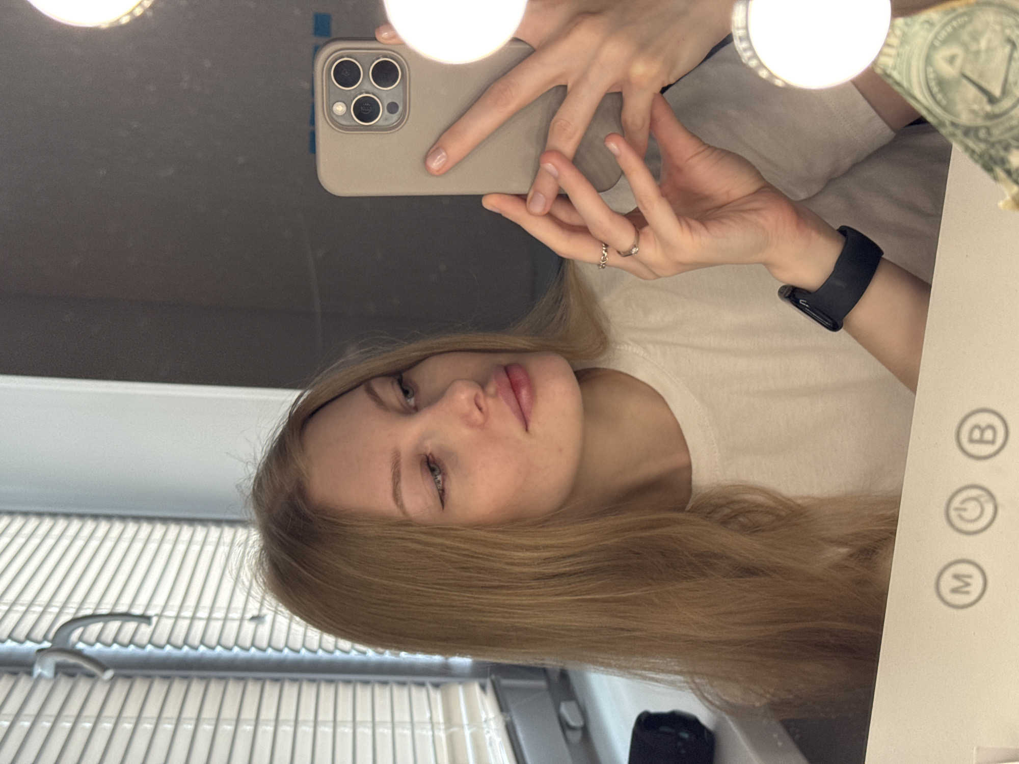 A young woman with long, blonde hair is taking a selfie in a bathroom mirror. She is holding a smartphone with a light beige case, showing its triple-lens camera. She is wearing a white t-shirt and a black smartwatch. A vanity mirror with lights around the edges is visible, and a US dollar bill is visible on the upper right side of the frame. The blinds are visible in the background and a bathroom sink is visible below. It is not possible to determine the location as Perm, Russia from the image alone.
