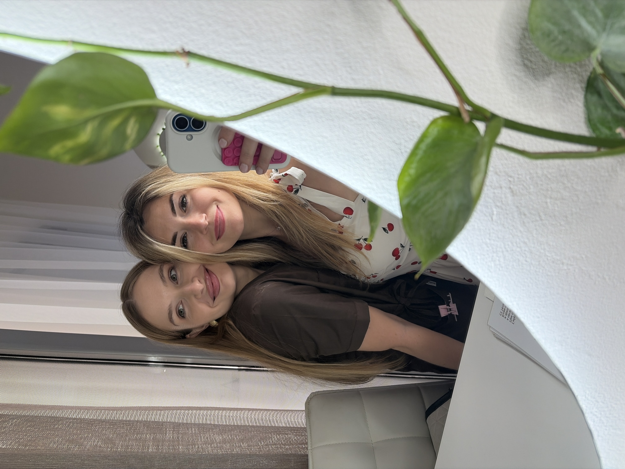 An indoor mirror selfie captures two young adult females in Budva, Montenegro. A prominent green vining plant in the immediate foreground partially obscures the lower right quadrant of the reflective surface.
The woman positioned on the left side of the mirror's reflection has long, light brown hair and wears a dark brown, short-sleeved top with a tie closure. She displays a soft smile, looking towards the right, and wears small gold hoop earrings. A small pink plastic clip is attached to her top. The woman on the right side of the reflection, holding the camera, has long, blonde hair and wears a white top featuring red cherry motifs and shoulder ties. She smiles directly at the camera, holding a white smartphone with a dual-lens camera system and a pink textured grip accessory in her right hand. Both individuals have rosy-pink lips.
The background behind the left woman reveals a window covered by light-colored, vertically striped sheer curtains. Behind the right woman is an unadorned white wall. A white quilted chair or headboard is partially visible in the lower left of the reflection. On a white tabletop surface in the lower center, a stack of white papers with printed text is present. The environment is brightly lit, indicative of natural light.