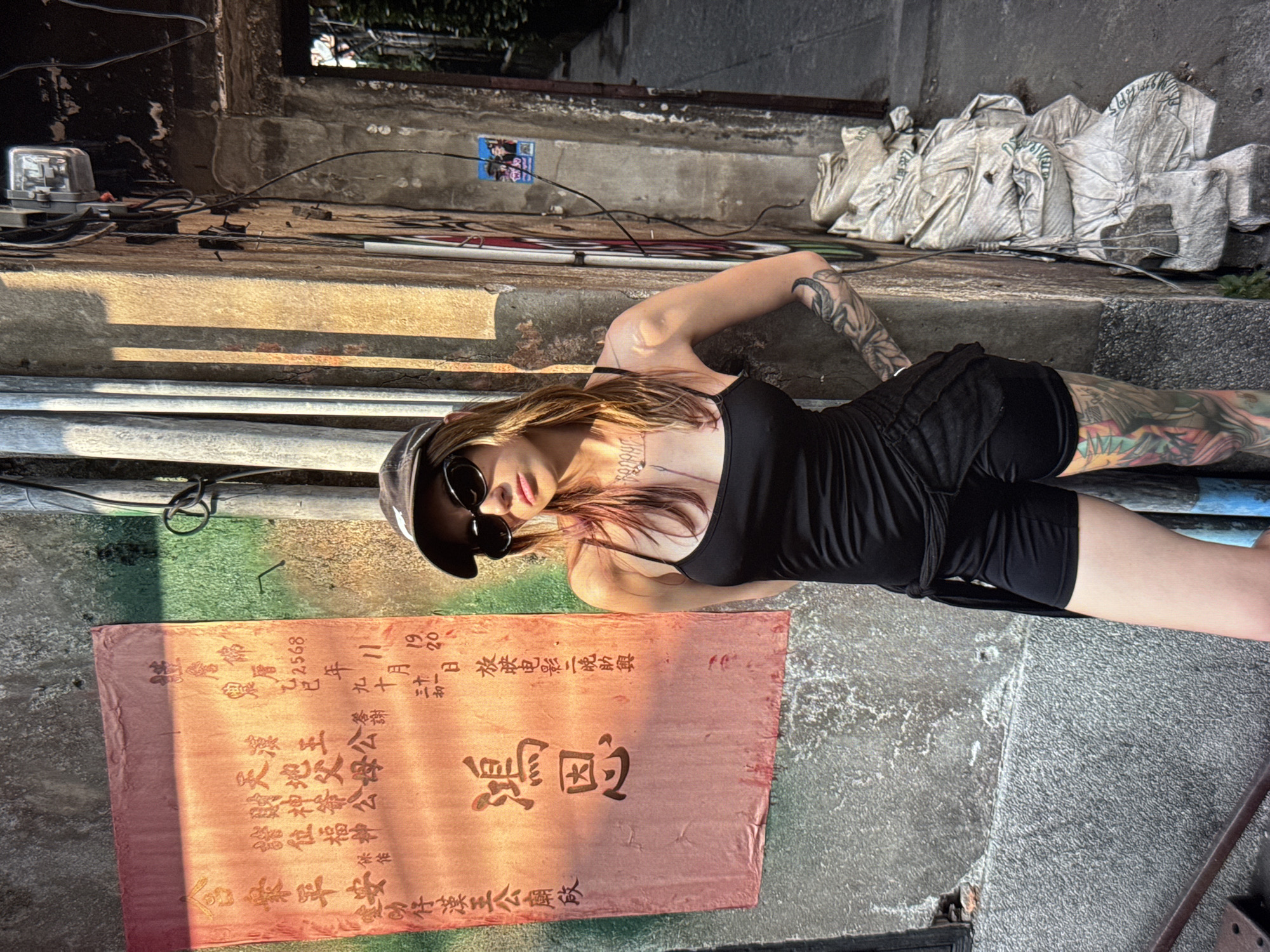 A young woman with blonde hair and a black baseball cap stands in front of a textured concrete wall. She is wearing a black tank top and black shorts. Her left arm is visible, featuring intricate tattoos. Her right arm is bent, with her hand resting on her hip. Behind her, a vertical piece of orange fabric with red Chinese characters is attached to the wall. The wall is also adorned with pipes and electrical wires. To the right of the woman, a narrow alleyway is visible, with a stack of white sacks piled against the wall. The scene is illuminated by a warm, directional light that casts long shadows.