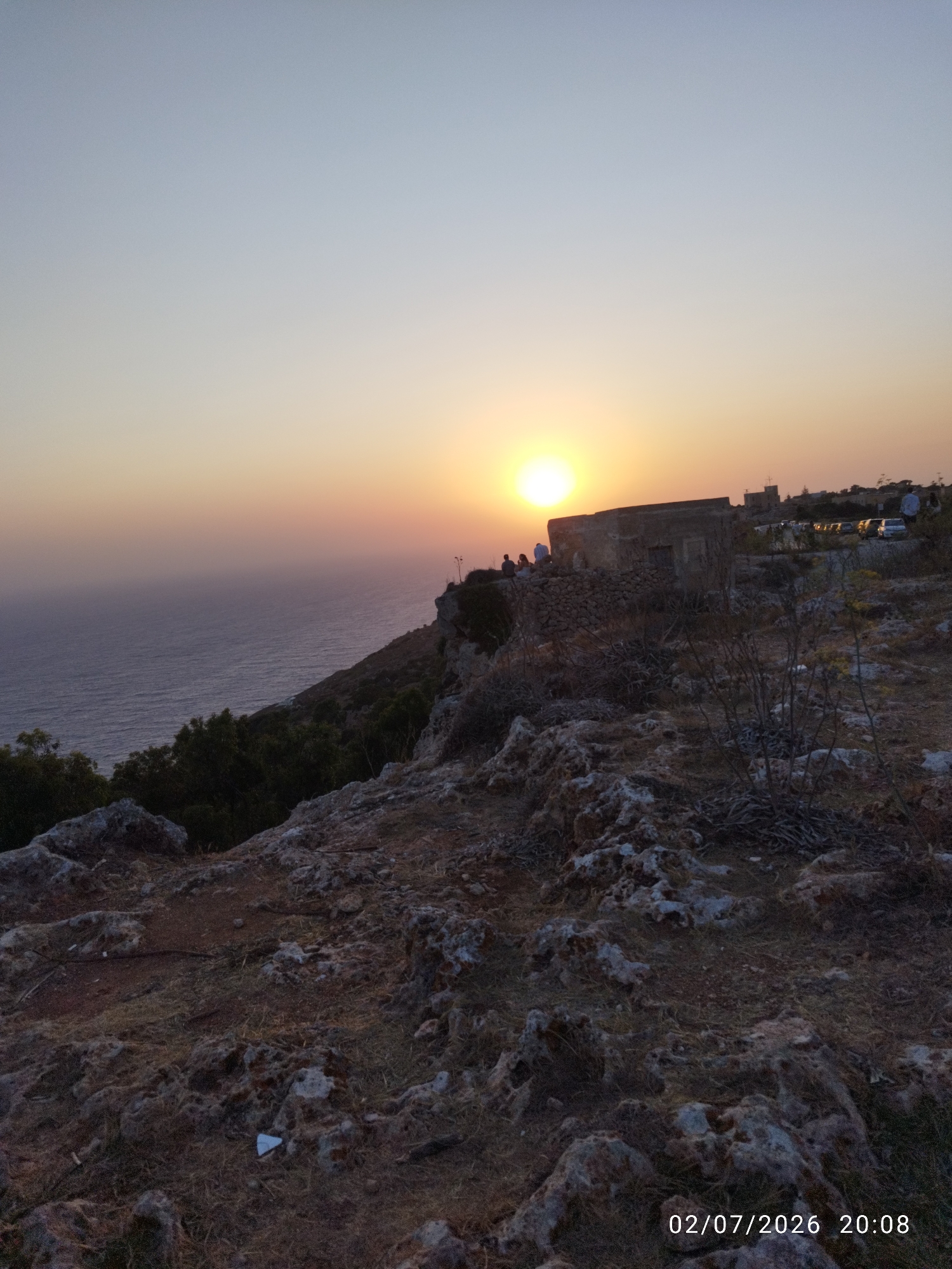 The image captures a sunset view from Dingli Cliffs, Malta. A vast expanse of the Mediterranean Sea stretches to the horizon, meeting a gradient sky of pale blue to soft orange. The rugged, rocky terrain of the cliffs dominates the foreground, sparsely covered with dry vegetation and small stones. On the cliff edge, a few people are visible, some seated and others standing, observing the sunset. Behind them, a weathered, stone building with a simple, unadorned facade stands prominently. Further in the background, several vehicles are parked, suggesting a popular viewpoint. The overall scene is tranquil, bathed in the warm, fading light of the setting sun.