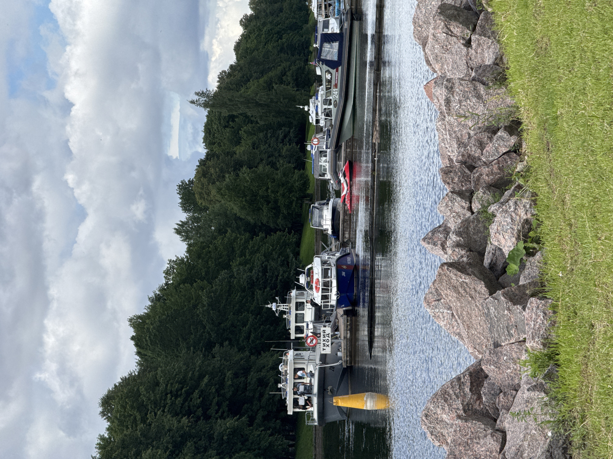 A fleet of patrol boats is docked at a harbor on a cloudy day. The boats are various sizes and colors, with some featuring red and blue accents. Several individuals are visible on the decks of the boats, appearing to be crew members or personnel. A yellow buoy floats in the water near the boats. In the background, a dense line of green trees forms a natural backdrop against the sky. The foreground is occupied by a rough shoreline composed of large, pinkish-grey granite rocks, with a strip of green grass. This scene is located at the Kronstadt Naval base, on Kotlin Island, in Saint Petersburg, Russia.