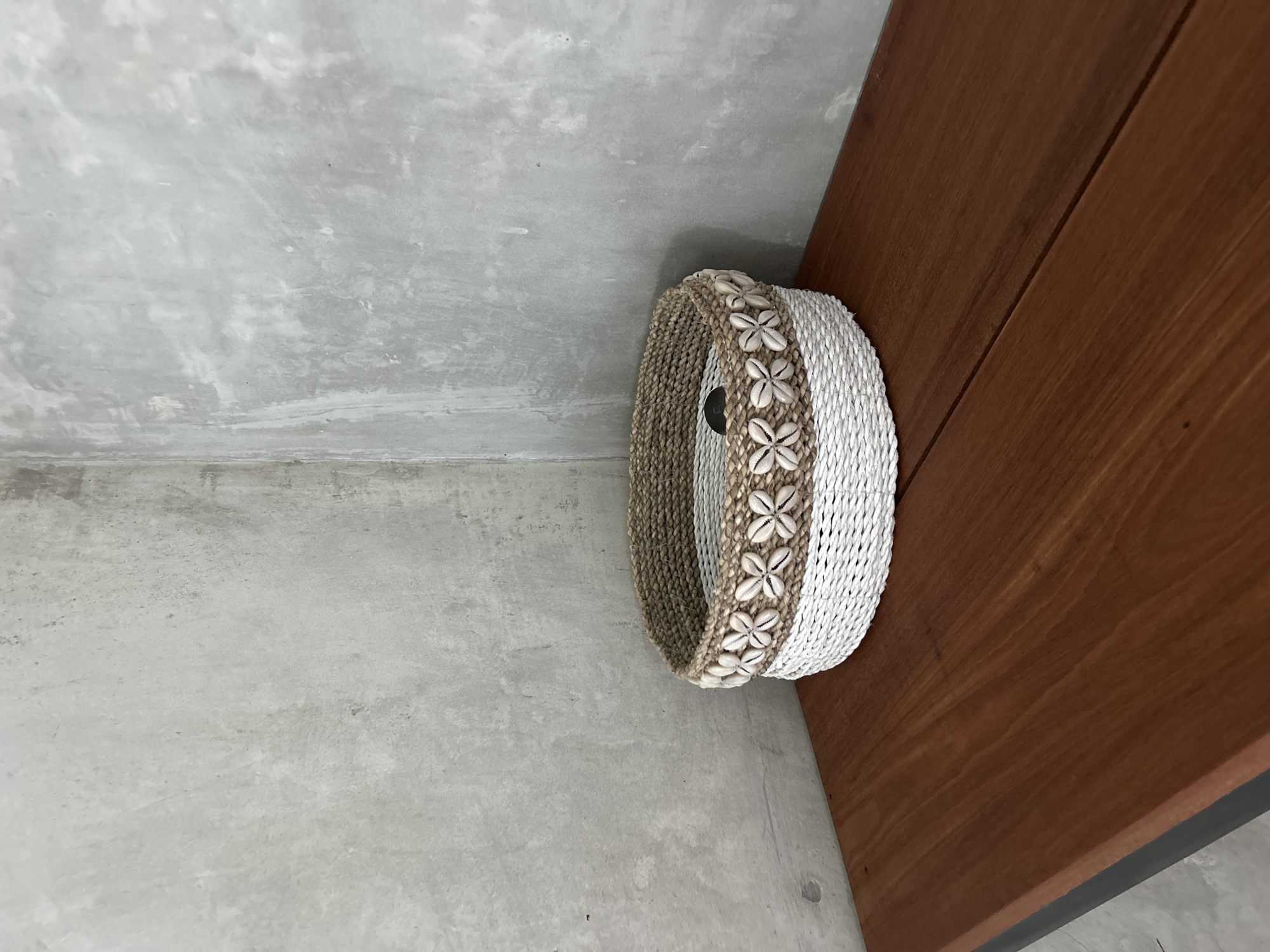 A circular, woven basket with a white base and a natural fiber top, decorated with a band of small, white shells arranged in a floral pattern, sits on a polished, reddish-brown wooden shelf. The shelf is positioned against a textured, gray concrete wall that forms a corner. The basket is centered on the shelf and appears empty. The lighting is soft and diffused, casting gentle shadows. The overall scene suggests an interior space with a rustic and natural aesthetic. This scene is located in Tembuku, Indonesia.