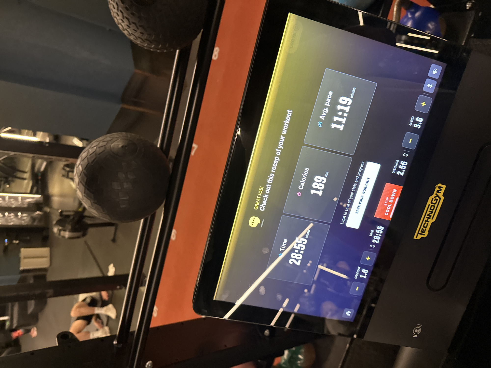 The image shows the display screen of a Technogym treadmill in a gym setting. The screen displays workout statistics: a time of 28:55, 189 kcal burned, an average pace of 11:19 min/km, distance covered of 2.56, and a speed of 3.6. A red stop/cool down button is visible. The environment includes other gym equipment, such as medicine balls on a rack. A person can be seen in the reflection of the treadmill screen. There is no evidence to specify the exact location in the city of London, United Kingdom.