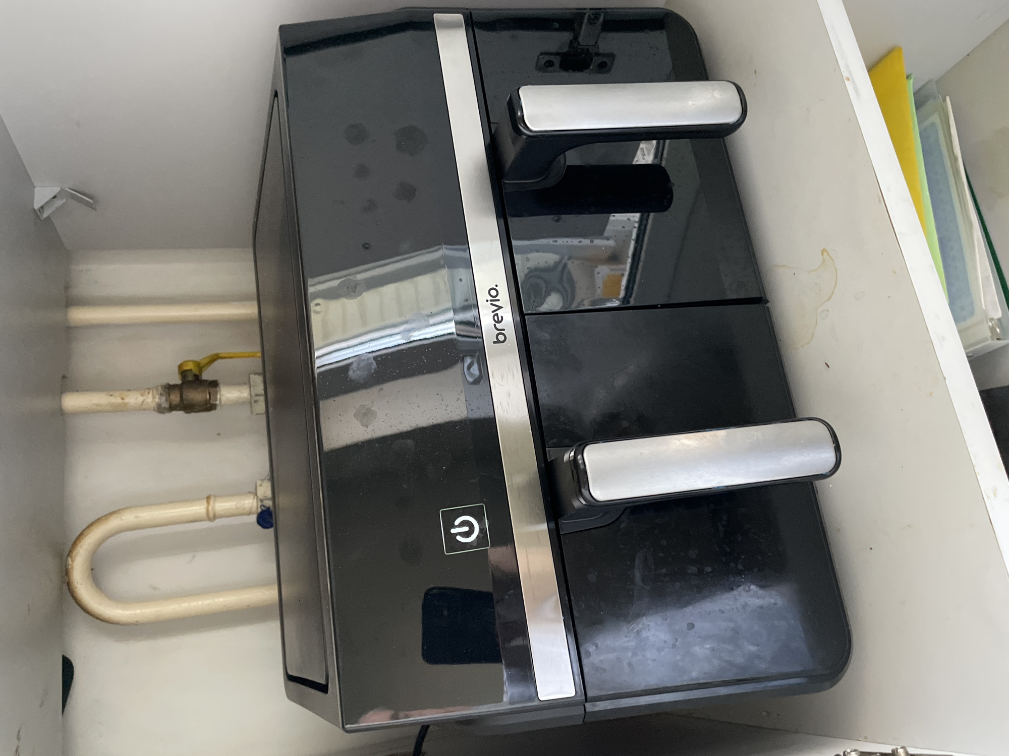 This is a close-up, eye-level, interior shot of a white kitchen cabinet. The cabinet contains a black Breville brand dual-basket air fryer. The air fryer is positioned centrally within the frame. Above the air fryer, a section of white plumbing pipes and a gas connection are visible. To the right of the air fryer, a small white bracket is affixed to the cabinet wall. On the shelf below the air fryer, several folders are stacked, a yellow one on top, followed by blue and green ones. A light-colored liquid stain is visible on the shelf surface to the left of the folders. The overall lighting is bright, with some reflections on the shiny surface of the air fryer, suggesting it is a well-lit kitchen environment. The image is taken from the perspective of someone standing and looking into the cabinet.