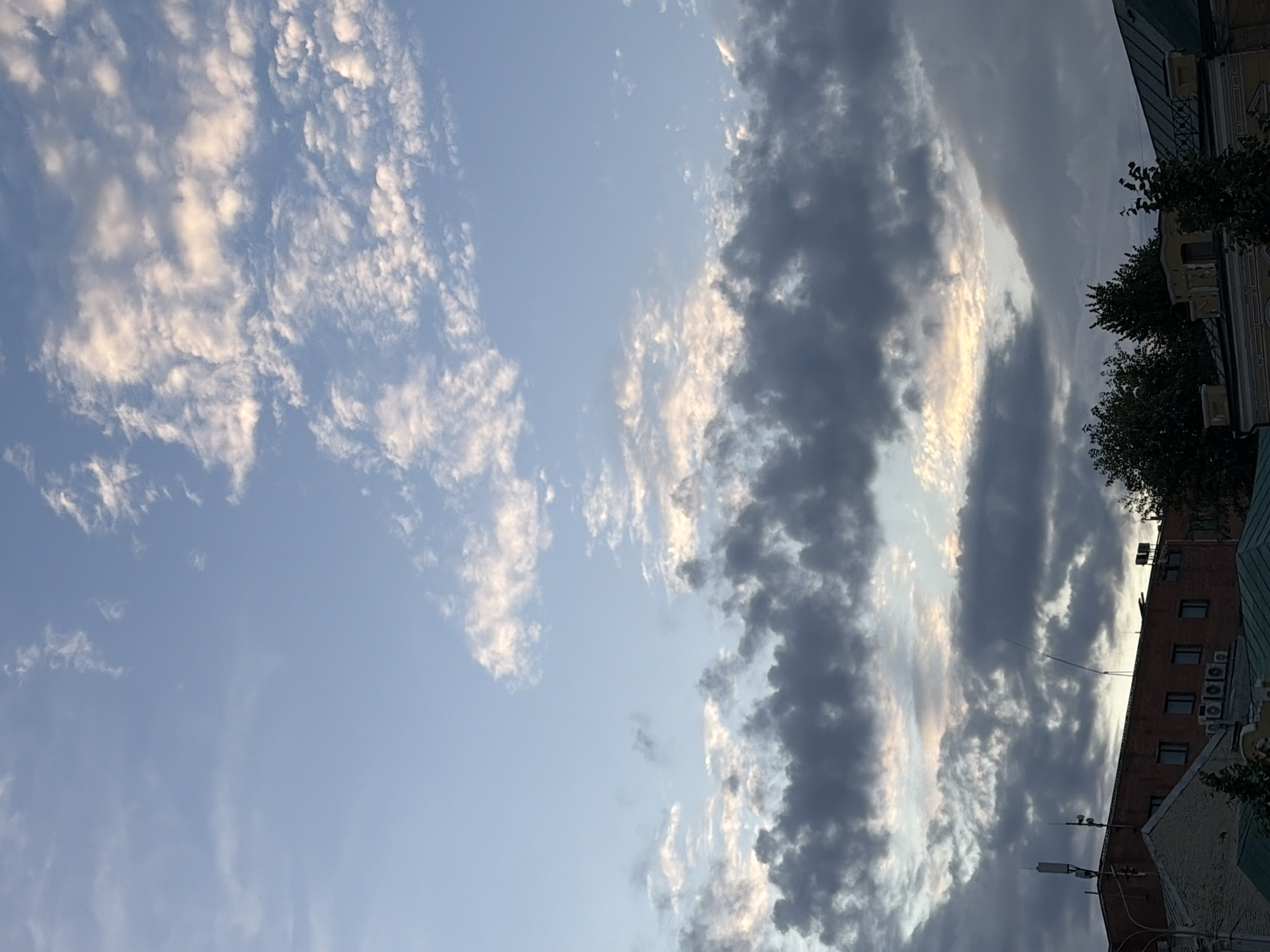 This is a vertical, eye-level medium shot of a cloudy sky above buildings in an urban environment. The sky is a pale blue with scattered white and gray clouds. Some of the clouds are illuminated by the sun, creating a bright contrast against the darker clouds. Below the sky, the tops of several buildings are visible. One building is made of red brick with several windows. Another building is partially visible on the left and is made of white brick with a green roof. Trees are also visible in the foreground and background. The scene appears to be an overcast day, possibly in the late afternoon or early morning.