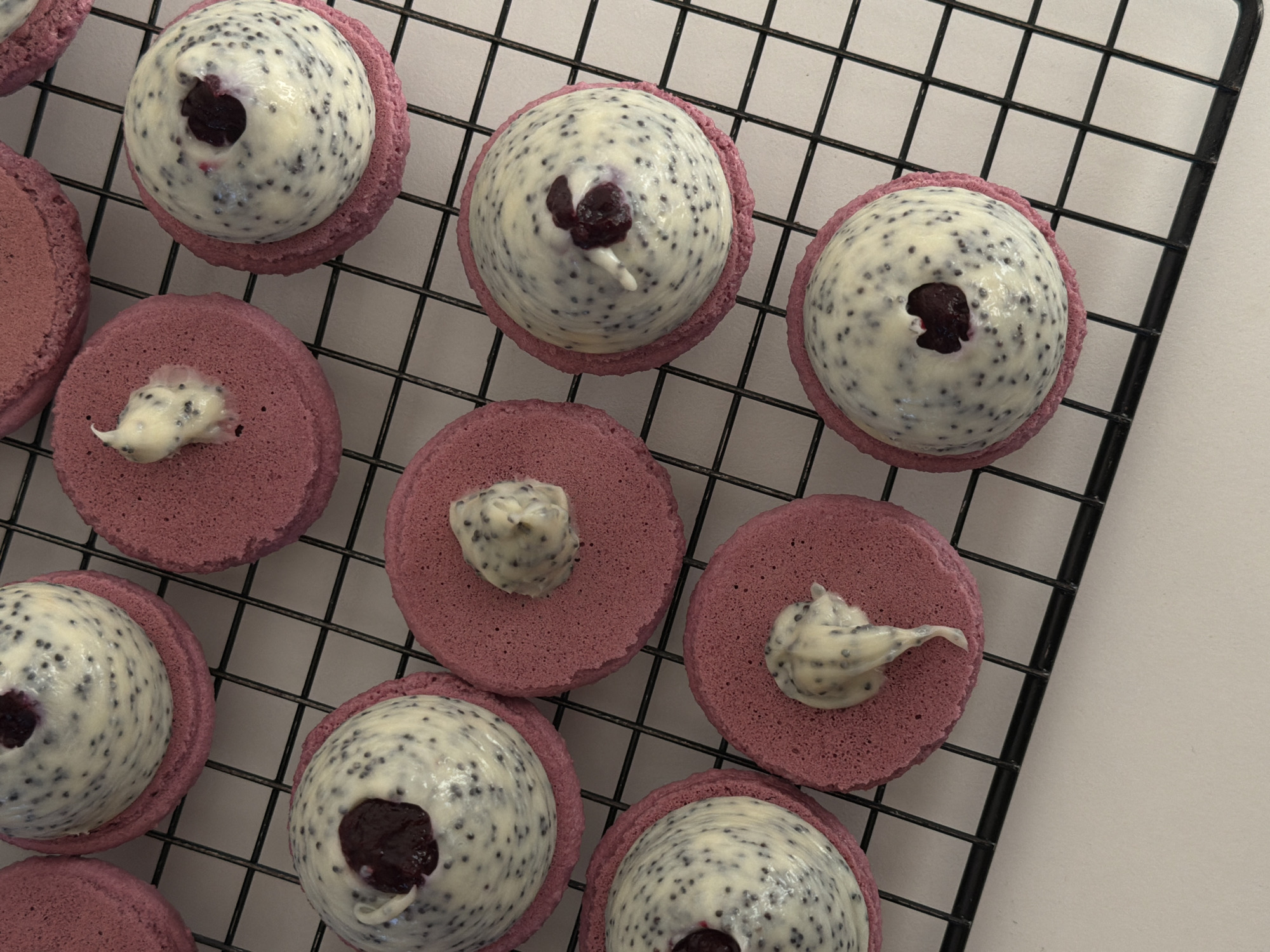 The image shows several macarons of a pinkish-purple hue arranged on a black wire cooling rack against a light, possibly white, surface. Some of the macarons are topped with a large, dome-shaped filling that is white with black specks. A few of these filled macarons have a dark, possibly fruit-based, center. Other macarons on the rack have a smaller dollop of filling. The macarons are closely packed together, partially obscuring the cooling rack beneath them. The setting appears to be indoors, possibly in a kitchen or bakery. The location, Tel-Aviv, Israel, cannot be confirmed from the image.