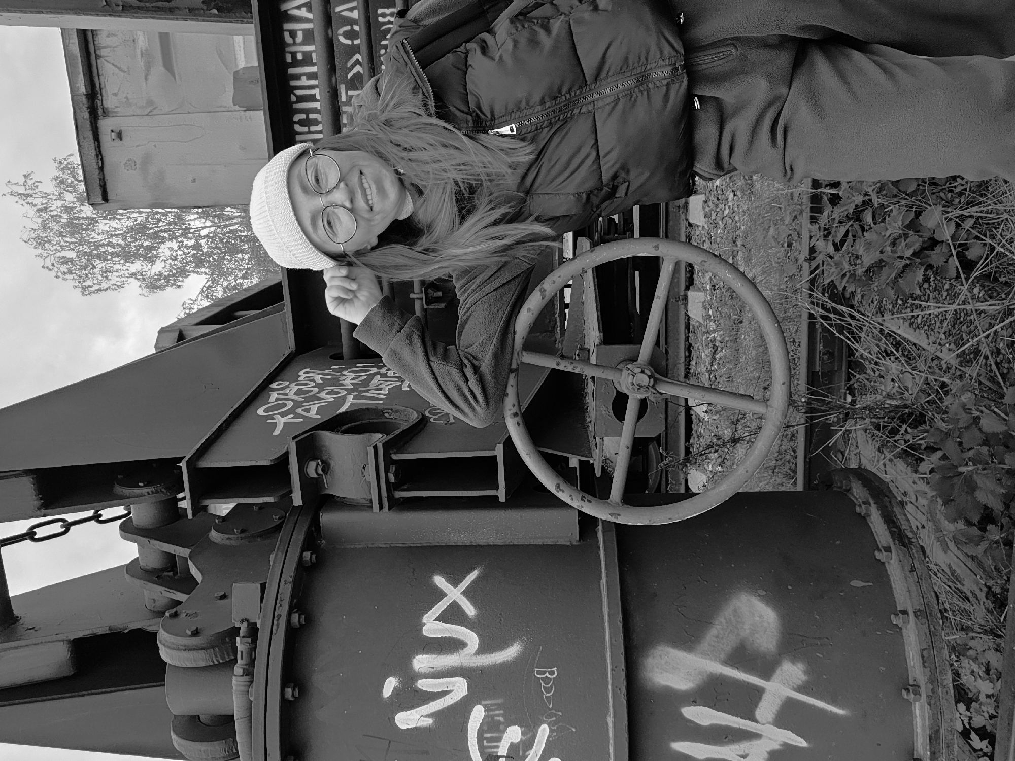A grayscale, slightly tilted, full-length shot of a woman in Chaikovsky, Russia, posing with a large industrial mechanism covered in graffiti.
The woman, seen from the chest up, wears a white beanie, round glasses, a dark puffer jacket, and a sweater. She smiles while holding the beanie's brim. Her long hair falls over her shoulder.
Behind her, a large, dark, presumably metal industrial object is visible. It has a large circular wheel, typical for operating heavy machinery. Graffiti in white paint covers the structure. The background consists of a building/container, a bare tree, and some vegetation. The location is in Chaikovsky, Russia.