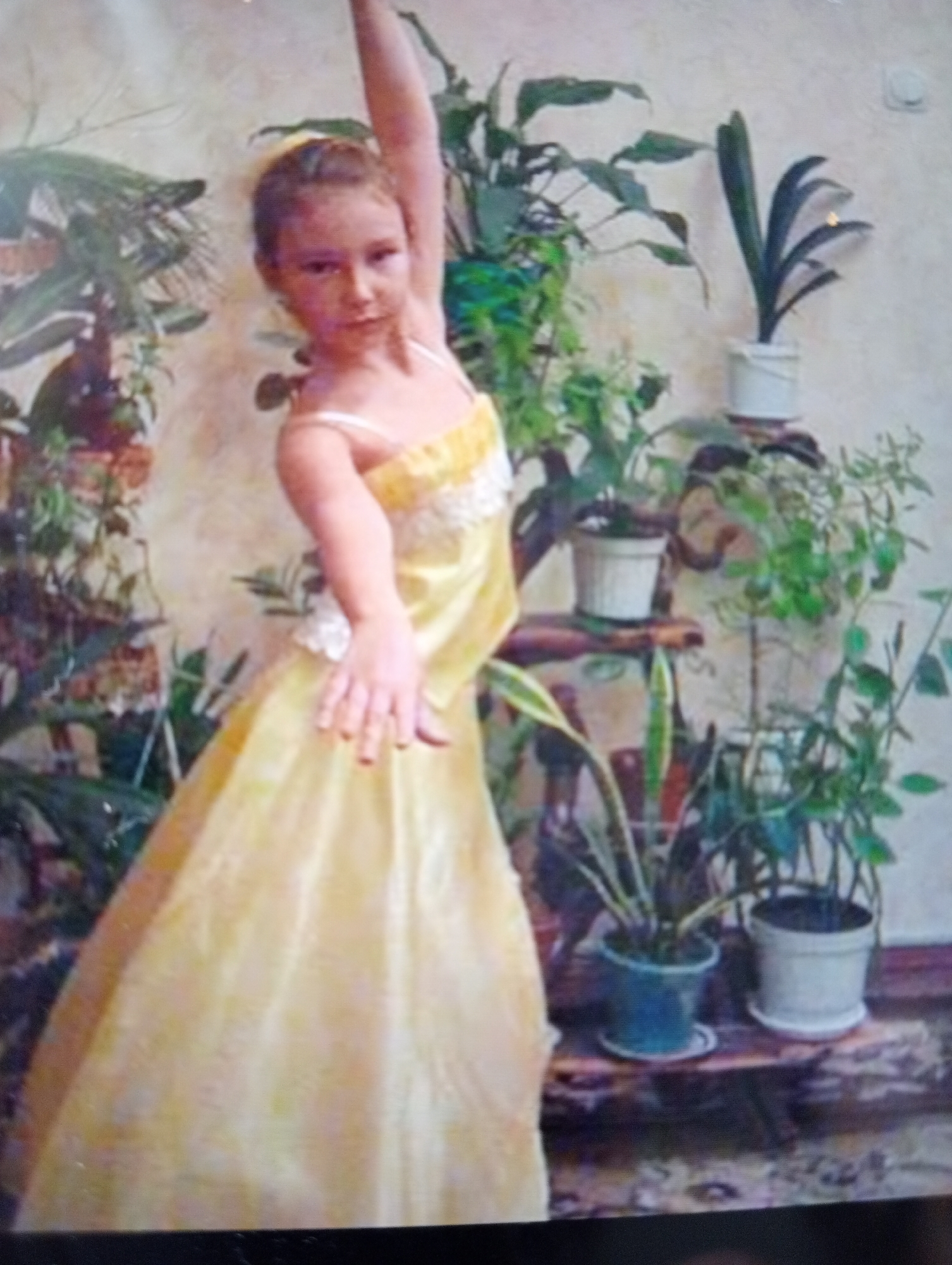 A young girl, appearing to be of East Asian descent, stands in a dance pose with her right arm extended upwards and her left arm bent and extended outwards. She is wearing a pale yellow, sleeveless dress with white lace detailing around the bodice and straps. Her hair is pulled back into a bun. The background is a textured wall with several potted plants. To the left, there are large, leafy plants. In the center and to the right, there are smaller plants in white and blue pots placed on shelves. The lighting appears to be artificial and the image is slightly blurry, suggesting it was taken with a low-resolution camera. The overall impression is of a domestic setting, possibly a home or a studio, where the girl is practicing dance.