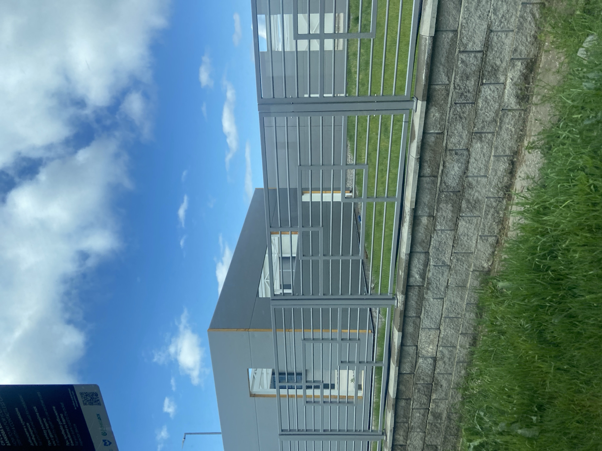 The image presents a partial view of a modern building in Zhiten, Bulgaria. The building features a gray exterior and rectangular window openings. A gray metal fence with horizontal bars runs parallel to the building, with grass visible between the fence and the structure. A retaining wall of textured, gray blocks borders the grassy area. The sky above is blue with scattered white clouds. To the bottom left, a sign with Bulgarian text is partially visible, indicating a contactless payment method.