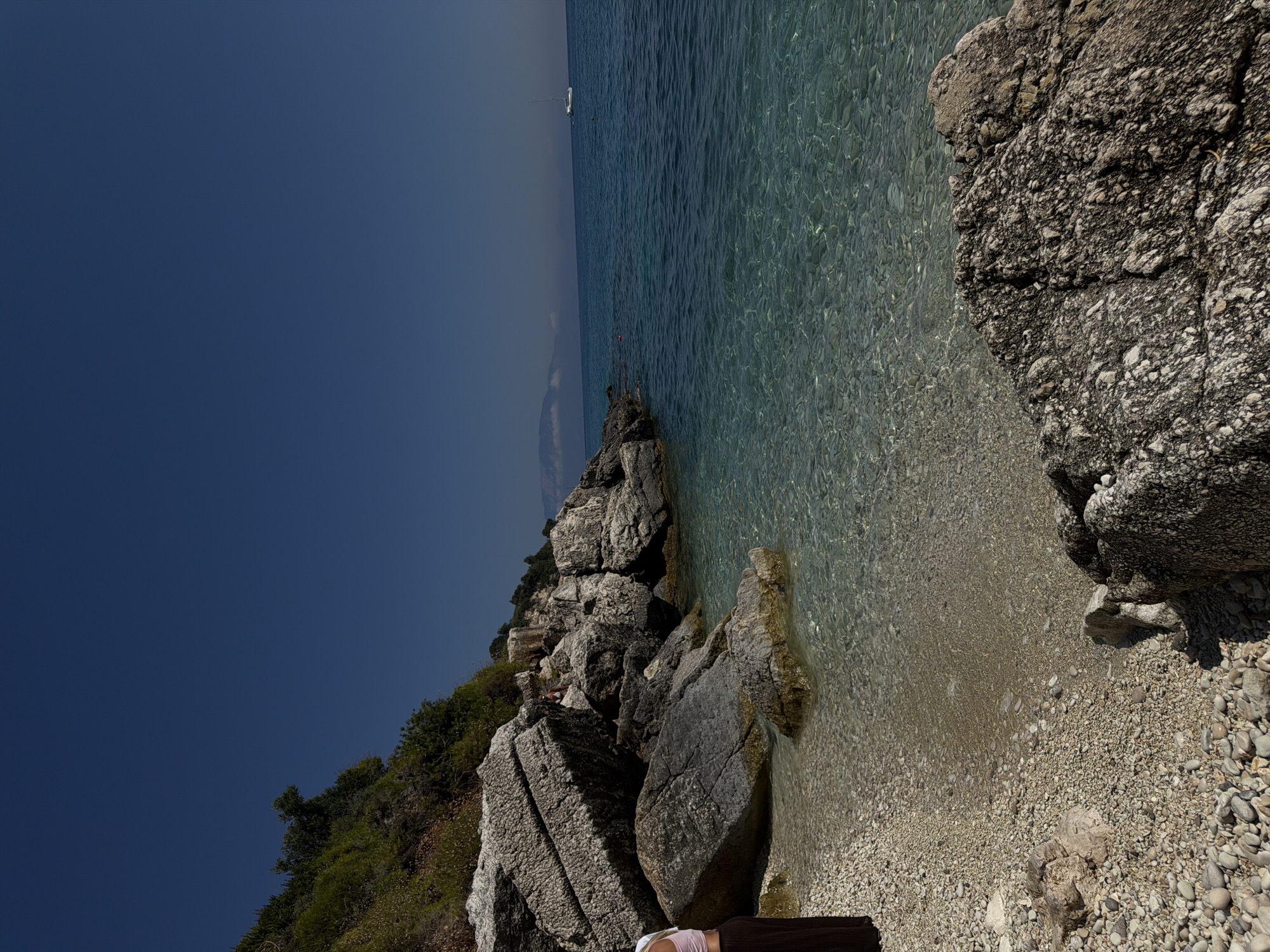 In a coastal scene, a person stands on a rocky beach under a clear blue sky. The beach is composed of small pebbles and larger rocks, with lush green vegetation covering the hillside that slopes down to the water. The ocean water is crystal clear, revealing the pebbly seabed beneath the surface. In the distance, a white sailboat glides across the turquoise sea, and a faint mountain range is visible on the horizon. The sunlight creates dappled patterns on the water and illuminates the scene with a bright, warm glow.