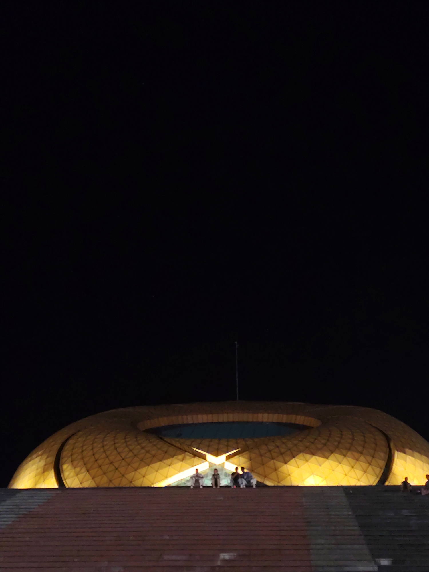 At night, the scene captures the iconic dome-shaped Cultural Palace of Da Lat, Vietnam. The golden exterior is illuminated, contrasting against the dark sky. Several people are sitting or standing on the stairs leading up to the palace. The foreground consists of the steps, and a pole extends upwards from the top of the dome.