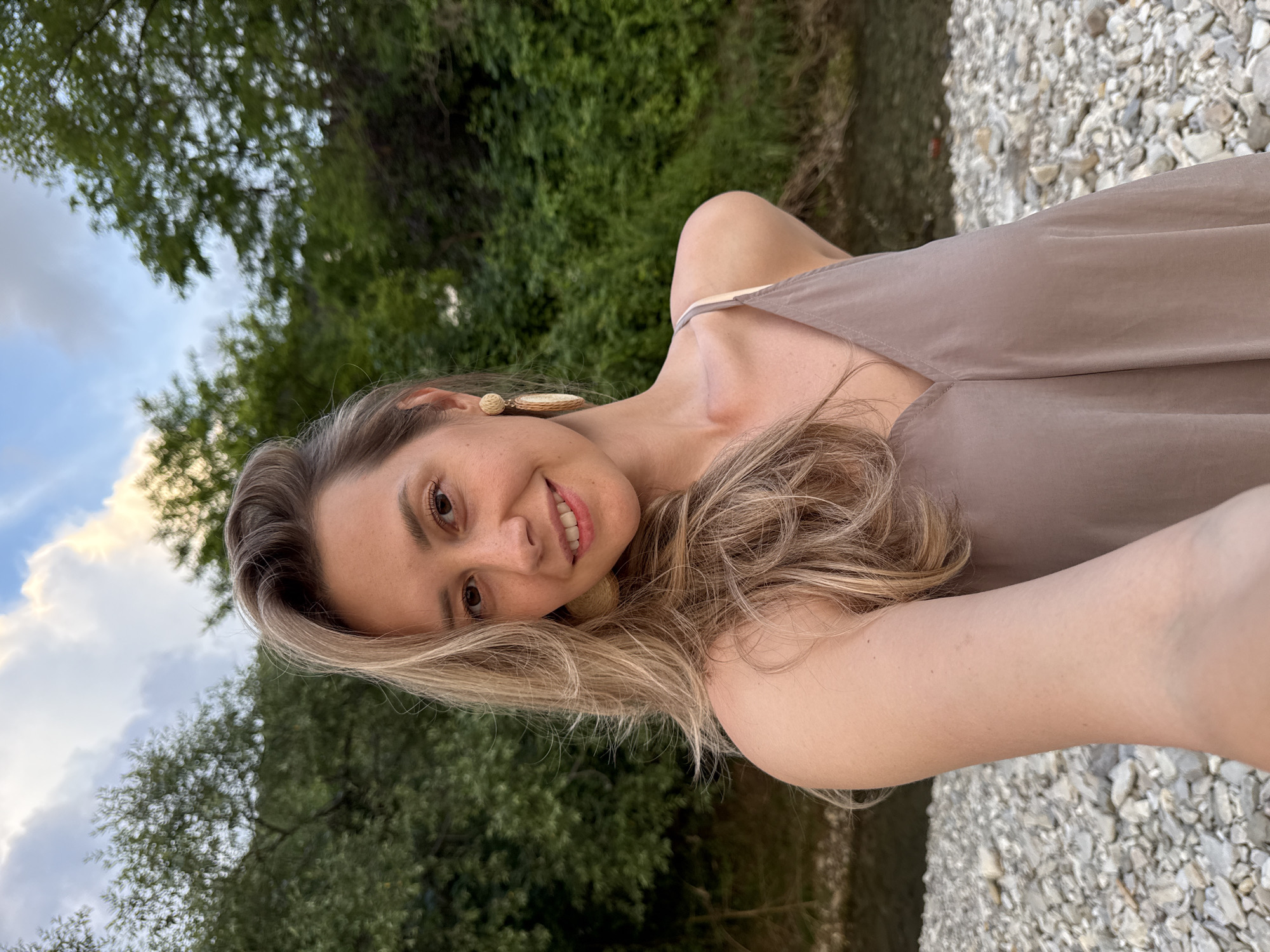 This media file is a selfie of a young woman with blonde hair, taken outdoors. She is smiling and looking at the camera. She is wearing a light brown dress with thin straps and a light-colored earring. The background shows lush green trees and a rocky area that appears to be a riverbed. The sky is visible in the upper left portion of the image, with some clouds and a bright light suggesting it is daytime, possibly late afternoon or early evening due to the lighting. The scene evokes a relaxed and natural setting. The image is vertically oriented, and the subject's head is tilted.