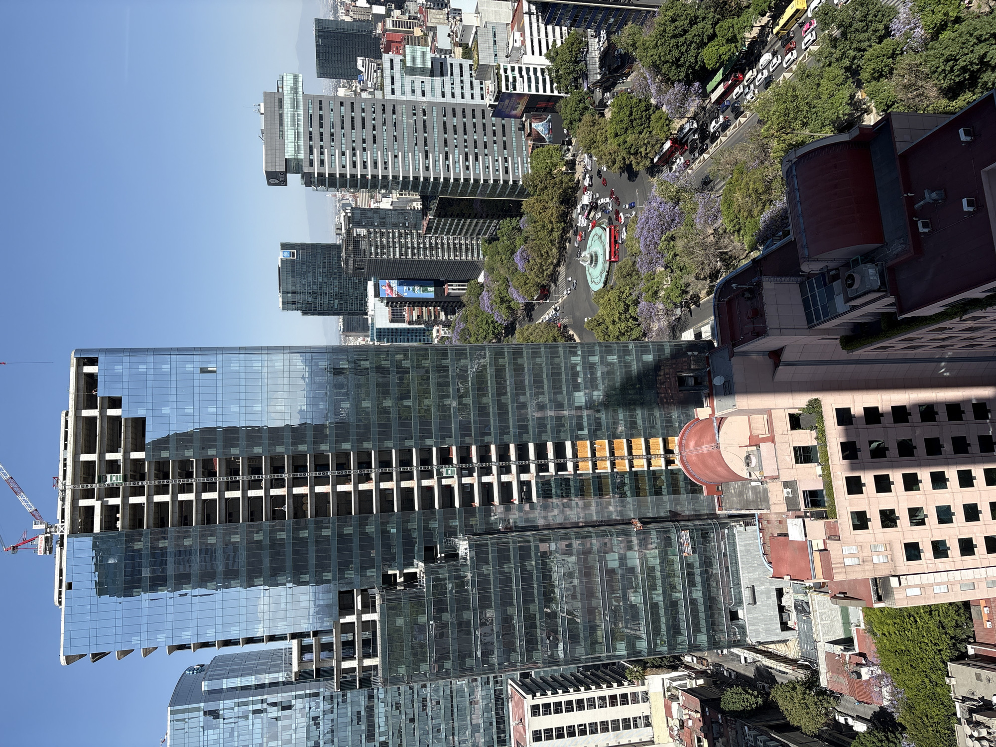An elevated forensic view depicts a bustling urban landscape in Mexico City, Mexico, centered on the Glorieta de la Diana Cazadora on Paseo de la Reforma. A large, modern skyscraper, partially clad with blue reflective glass and revealing exposed concrete structural frames on its upper levels, dominates the left frame. A red and white tower crane, labeled "ESPIEGA," is positioned atop this structure, indicating ongoing construction. A second, similarly styled glass high-rise, also under construction, is partially visible to the left. Below these buildings, a multi-lane circular roundabout contains a multi-tiered, light-colored fountain at its center. Heavy vehicular traffic, consisting of numerous passenger cars, multiple red double-decker tour buses, and other public transport vehicles, moves around the roundabout and along its connecting tree-lined avenues. Many trees display vibrant purple blooms, consistent with Jacaranda trees. To the right, a dense collection of diverse high-rise buildings, including one marked "SOFITEL," extends towards the horizon. In the lower right foreground, a multi-story building with a light-colored facade and a distinct reddish, rounded roof is visible. The sky is clear and light blue. A small scaffolding platform is present on the exterior of the primary skyscraper under construction, suggesting workers are present.
