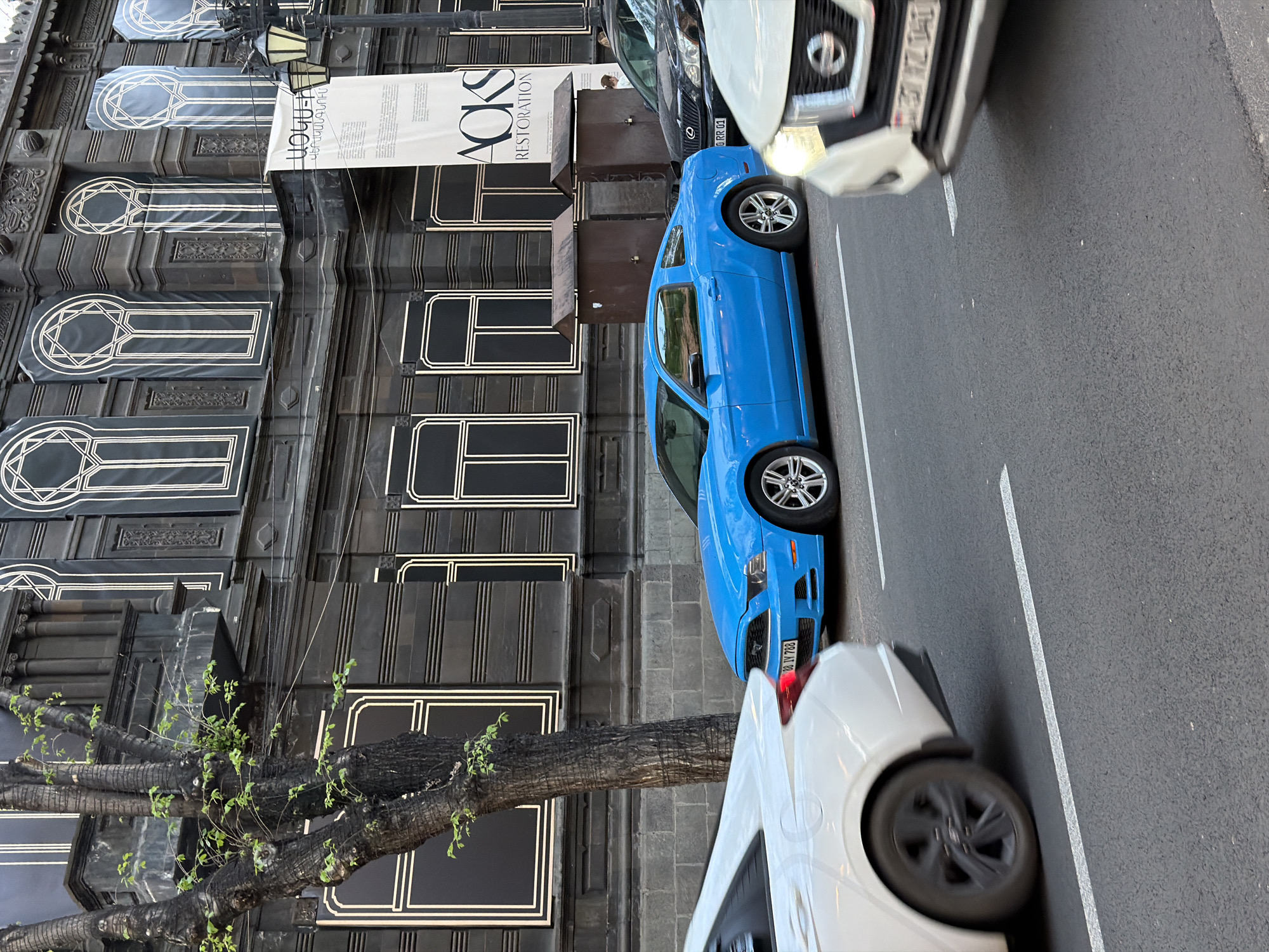 The image captures an urban street scene in Yerevan, Armenia, dominated by a large, dark-colored building with ornate architectural details. The building features numerous windows, some covered by tall, vertical banners. These banners prominently display "AOKS RESTORATION" and other text, including partially visible Armenian script. A person is faintly visible standing near the building's entrance, beneath one of the banners.
On the asphalt street, marked with white parking lines, several cars are parked. A bright blue Ford Mustang (license plate "08 VV 788") is positioned in the middle, facing the camera. Behind it, a dark Lexus SUV is visible. In the foreground, parts of two white cars are seen; one (possibly a Nissan SUV with "RR 01" on its plate) is on the far right, and another is closer to the bottom right.
A tree with thick, dark branches and sparse, light green foliage grows near the building on the left. The scene appears to be set during daytime with clear weather, reflecting a typical city environment. The visible text on the banners and the Armenian license plate format confirm the location context.