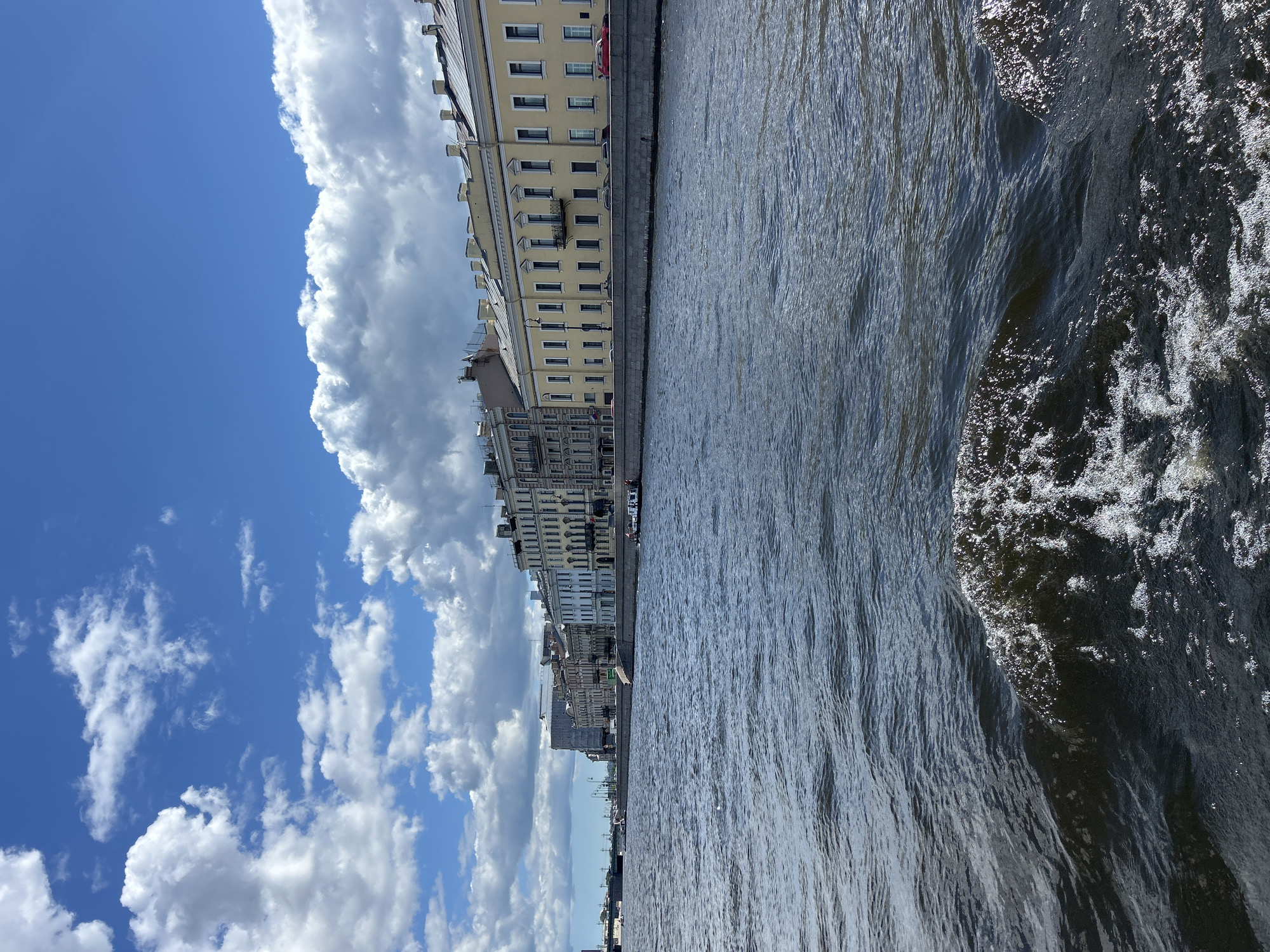 The image depicts a city skyline viewed from a body of water, likely a canal or river. The buildings are tall and multi-story, with a consistent architectural style featuring many windows and decorative elements. One prominent building is painted a pale yellow, while others appear to be in shades of grey and white. The architecture suggests a European city, possibly St. Petersburg, Russia, due to the classical design and the presence of a wide waterway.
The water itself is a dominant feature, with a rippled surface reflecting the sunlight and creating a sense of movement. On the right side of the image, the water is churning and frothy, indicating the presence of a boat or other vessel causing a wake. A few small boats are visible docked along the embankment.
The sky is a vibrant blue, filled with large, fluffy white clouds. The contrast between the bright sky and the dark water creates a dramatic visual effect. The sunlight suggests it is daytime, likely late morning or early afternoon.
No people are clearly visible in the image, though there might be tiny figures along the embankment or on the boats if one looks very closely. No text is visible in the image. The overall mood is serene and picturesque, capturing a classic urban waterfront scene on a bright, cloudy day.